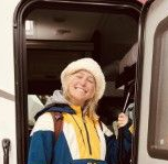 Woman wearing a bucket hat smiles while standing in a RV doorway; blue, yellow, and white jacket.