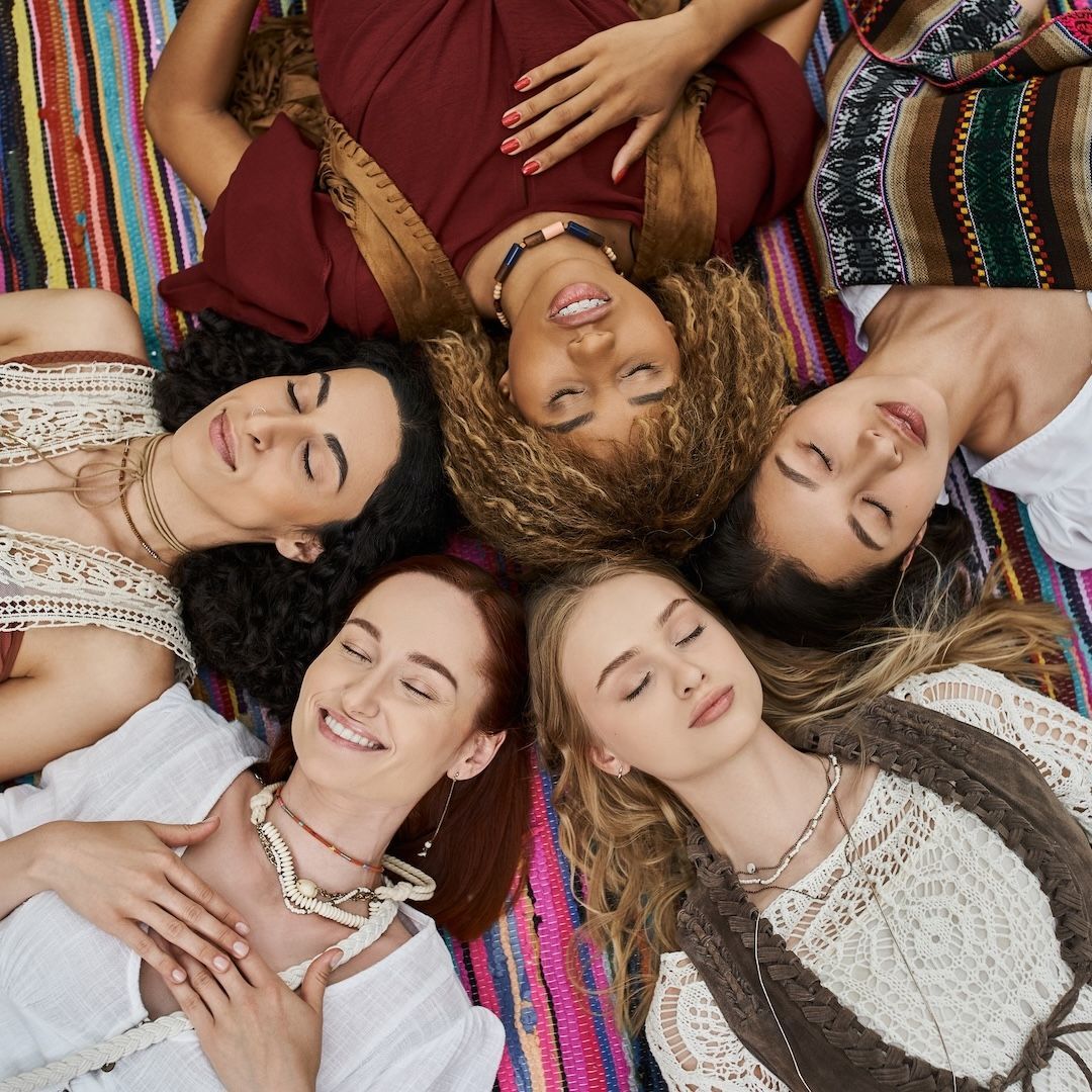 Five women lying in a circle on a colorful blanket, smiling, wearing necklaces, boho clothing.
