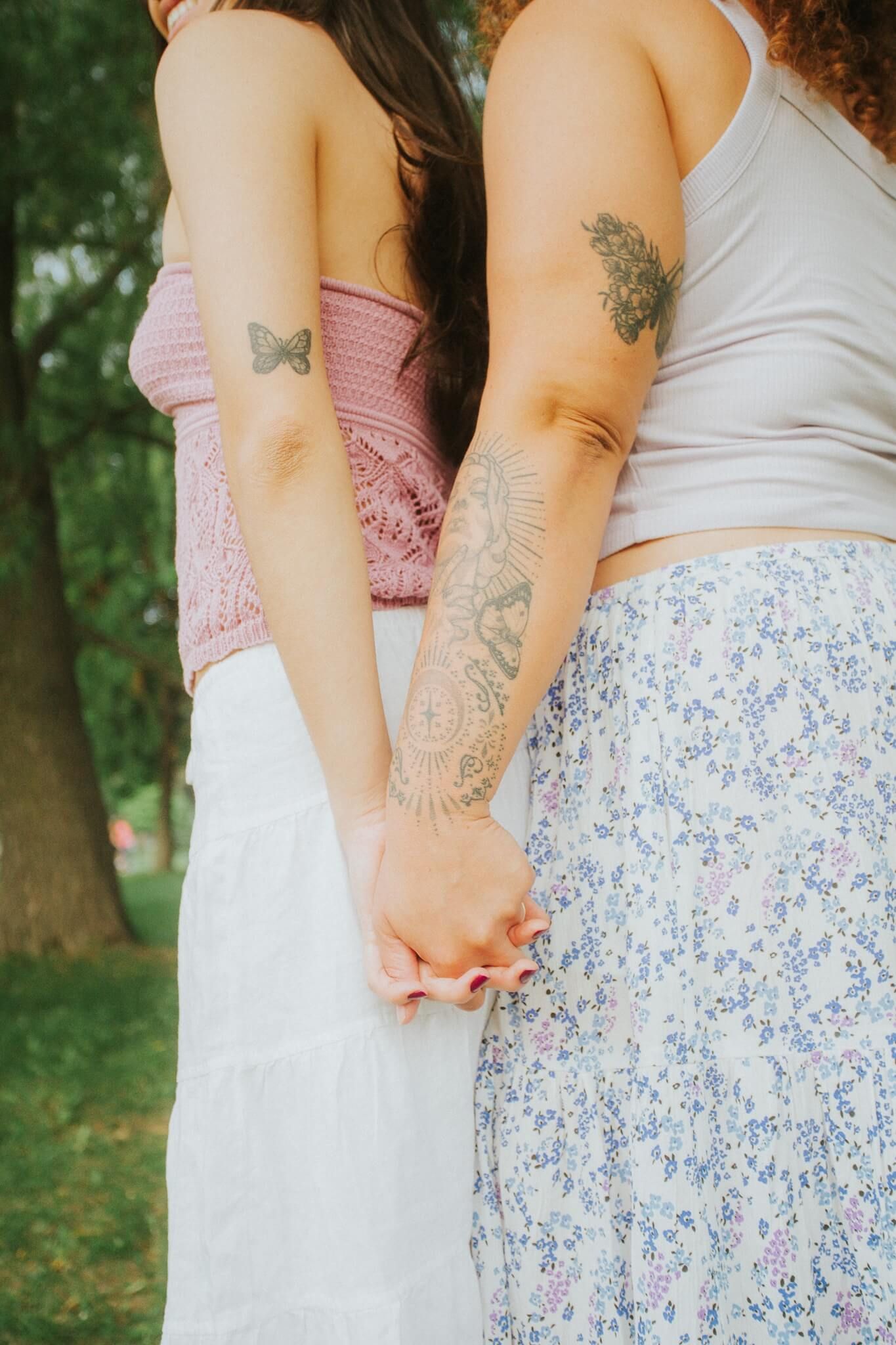 Two people holding hands, outdoors. One wears a pink top, the other a white top, both with arm tattoos.