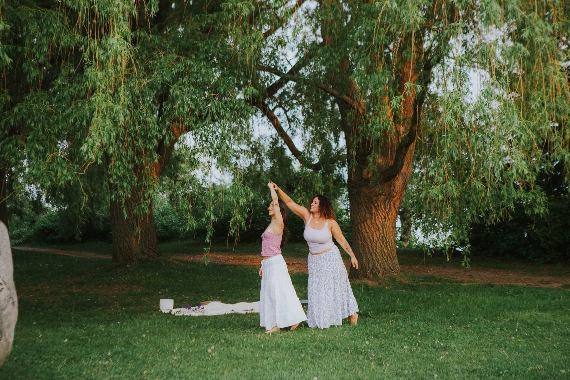 Two women dance under a willow tree in a park. One holds the other's hand, both in white skirts.