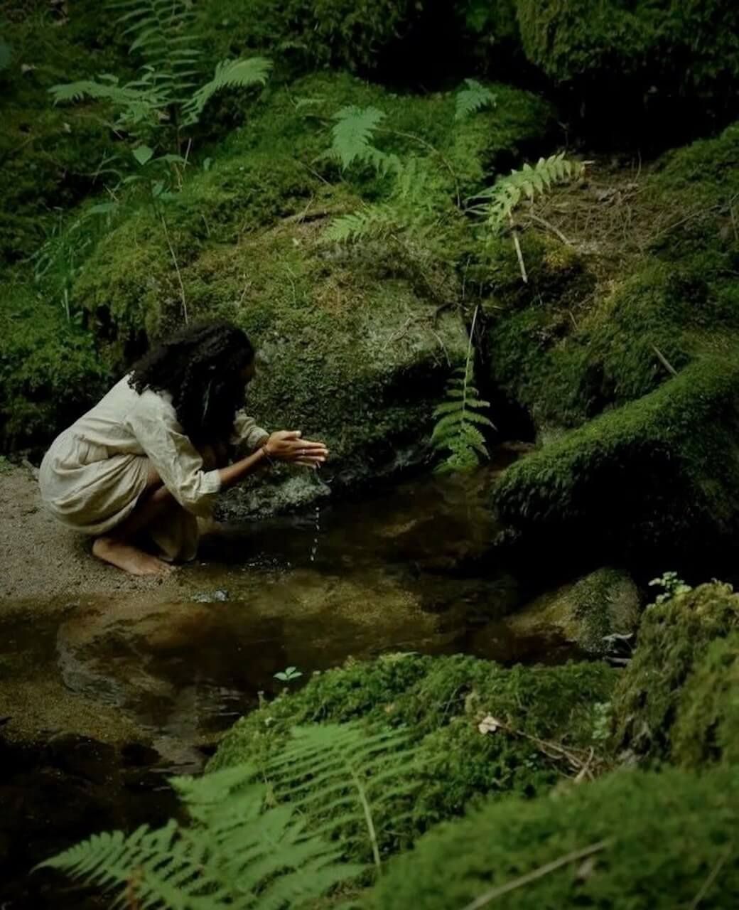 A woman crouched down with her feet planted in the sand and rubbing water between her hands in a forest