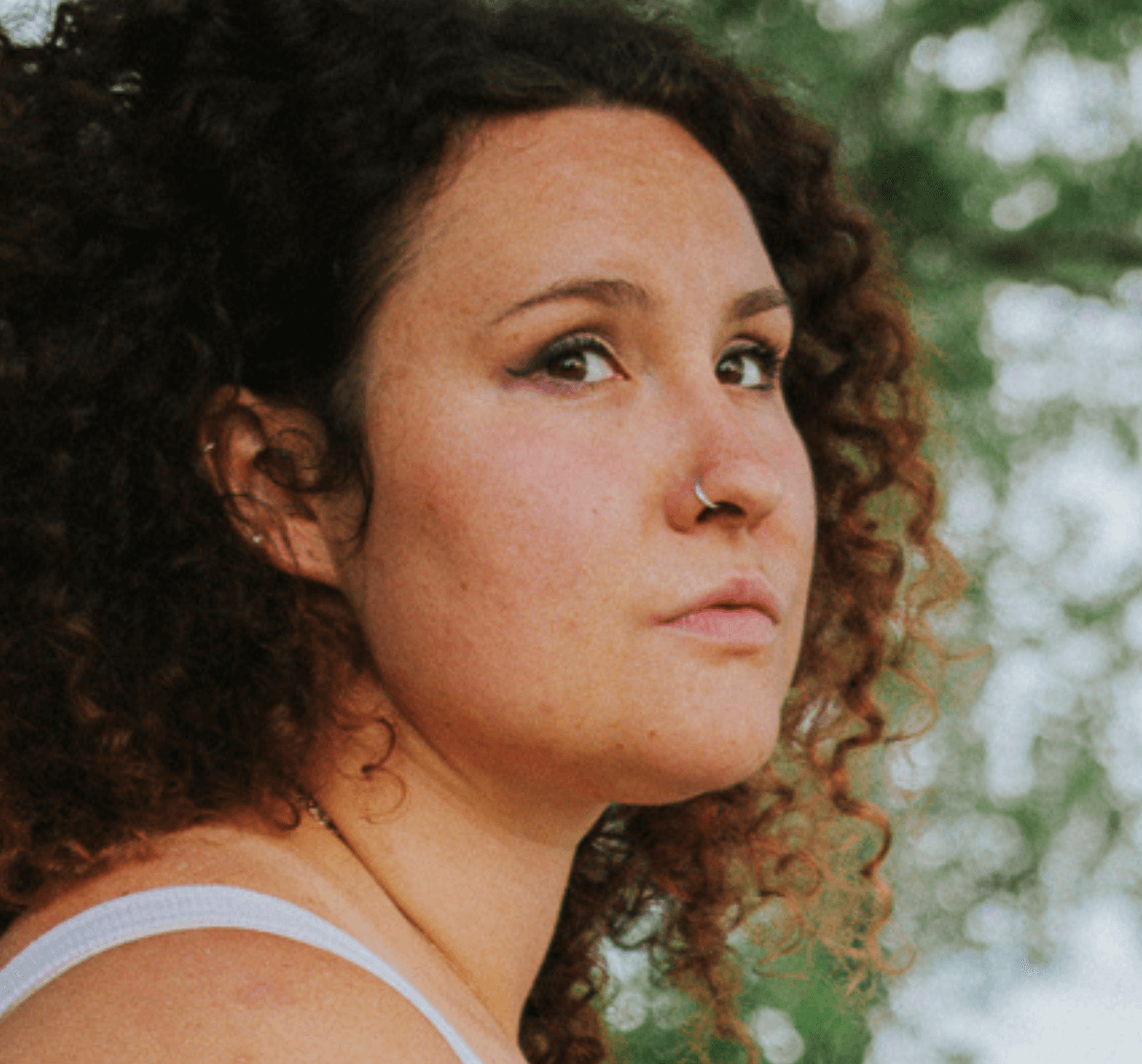 Woman with curly brown hair looks off-camera; a nose ring is visible. Outdoors, in front of blurred green foliage.