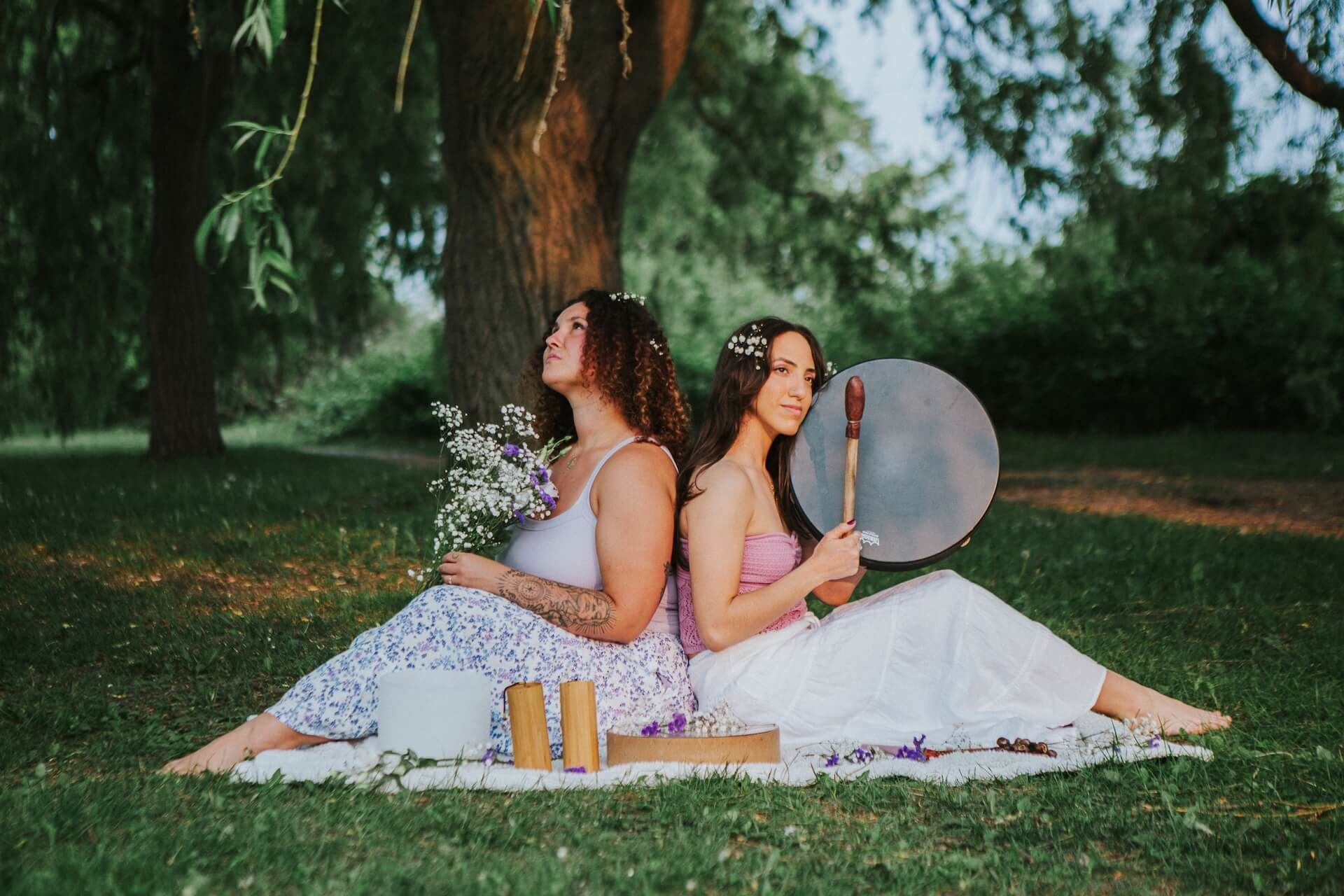 Two women sit back-to-back on a blanket in a park, holding instruments and flowers. Sunlight shines.