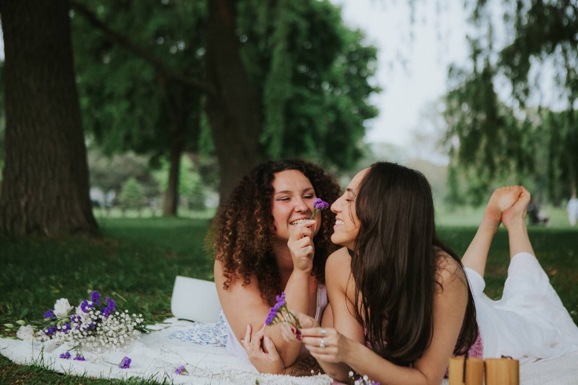 Two women smiling and laughing on a picnic blanket in a park, holding flowers.