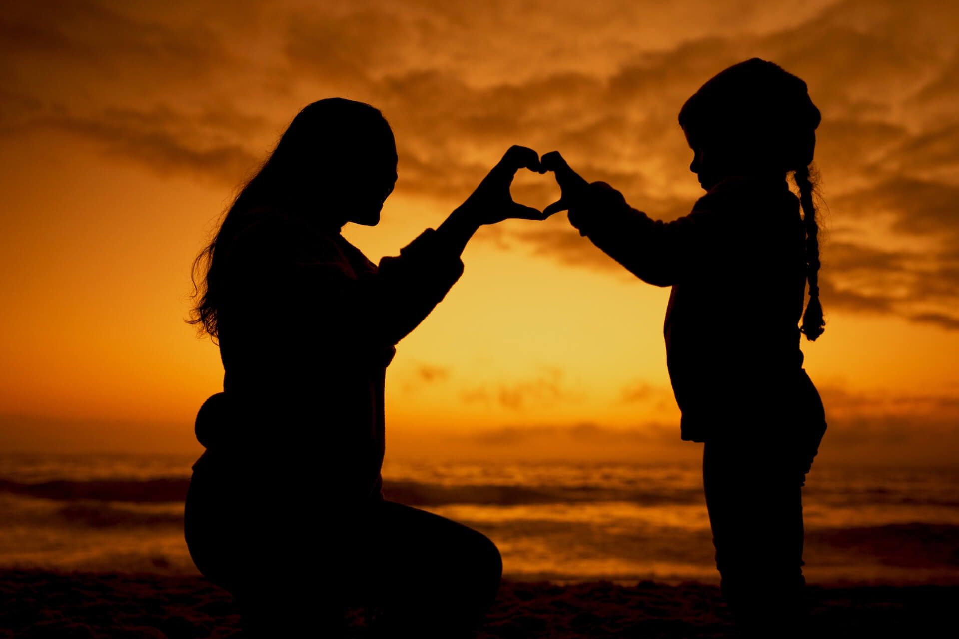 Silhouette of a person kneeling and a child making a heart shape with hands, against an orange sunset over water.