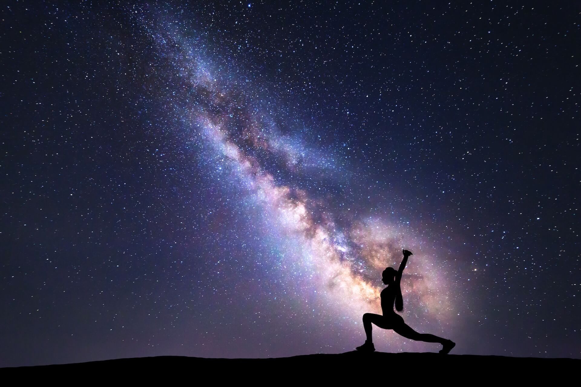 Silhouette of person in yoga pose, arm raised, under starry night sky of the Milky Way.