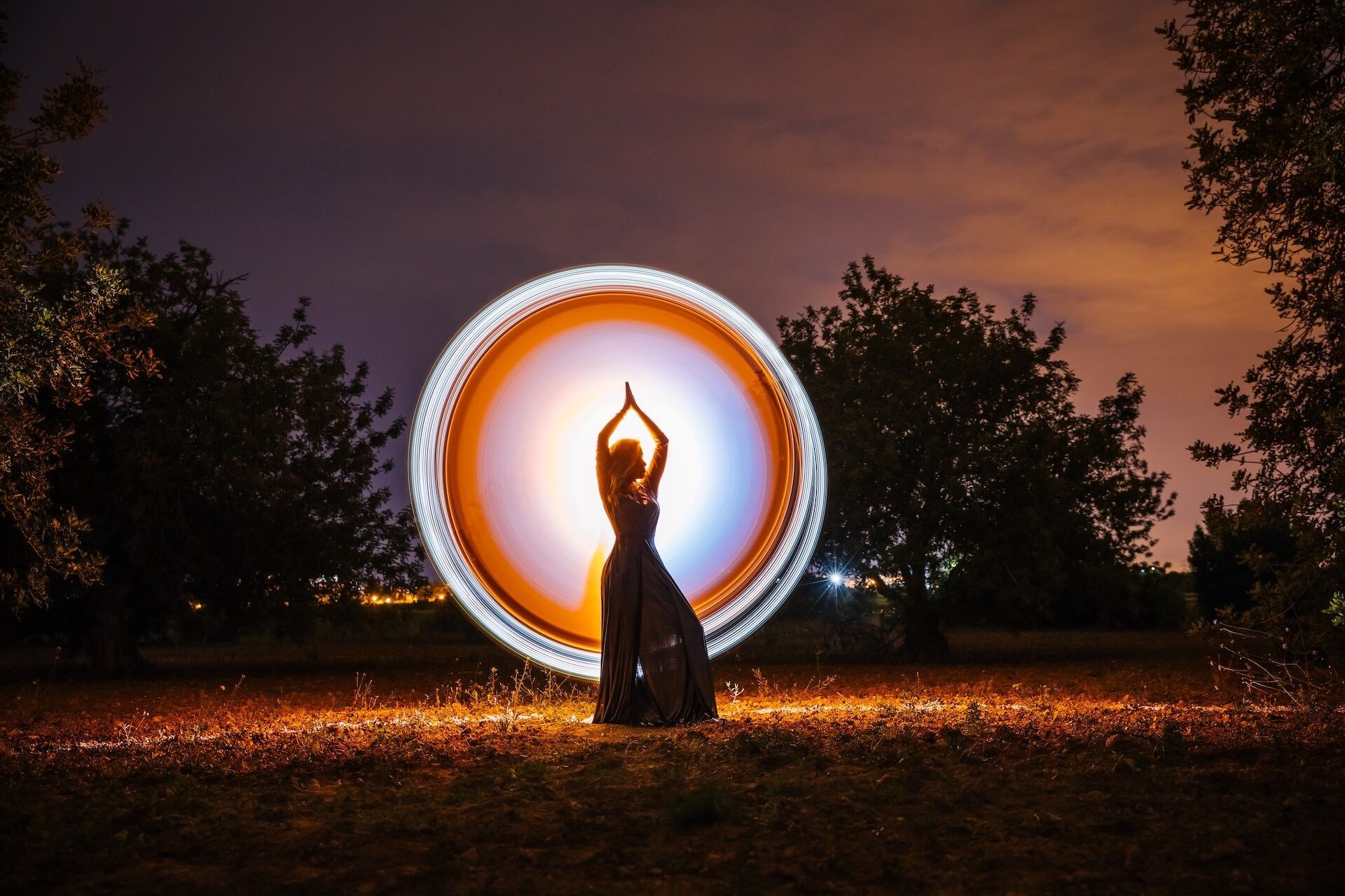 Silhouette of a person with arms raised inside a glowing circle of light in a park at night.