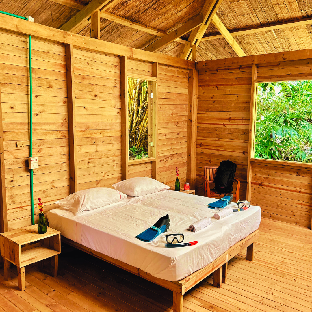 Wooden cabin bedroom with a double bed, snorkeling gear, and open windows with green outside.