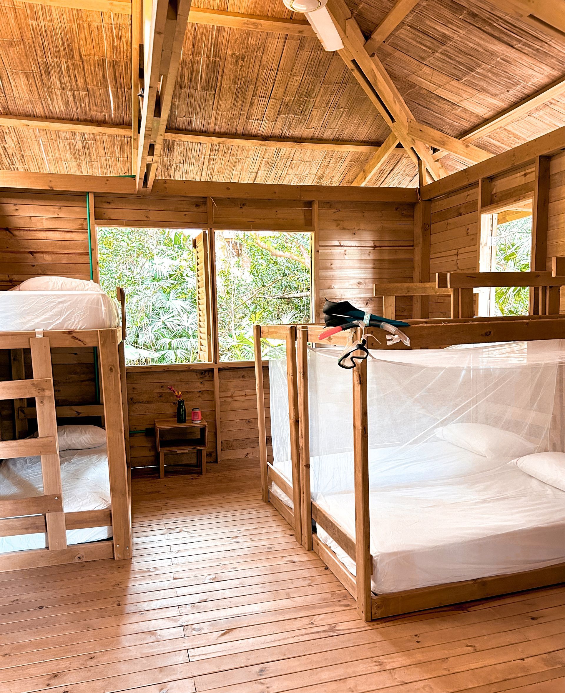 Interior of a rustic cabin with bunk beds, mosquito netting, and a window overlooking greenery. Wooden structure.