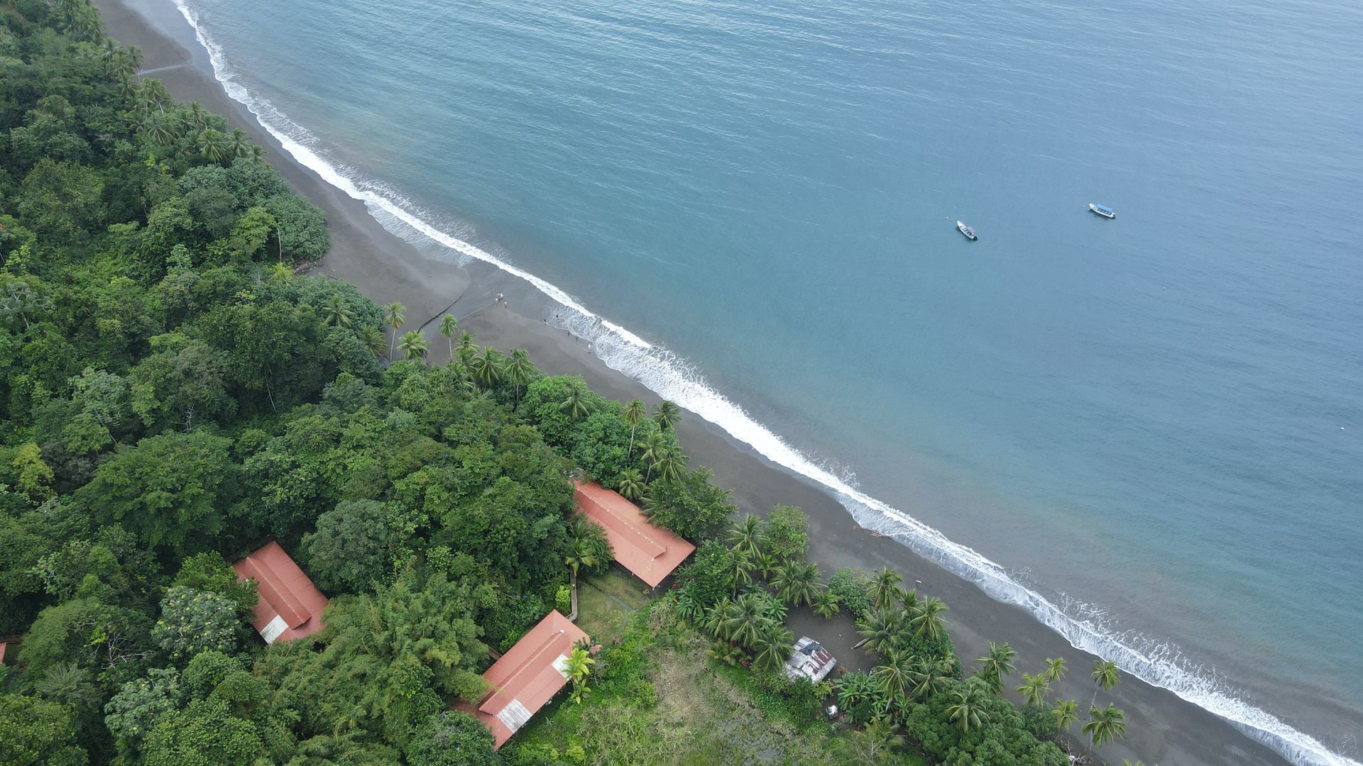Beach with cabins nestled in lush green trees next to dark blue ocean, three boats are on the water.