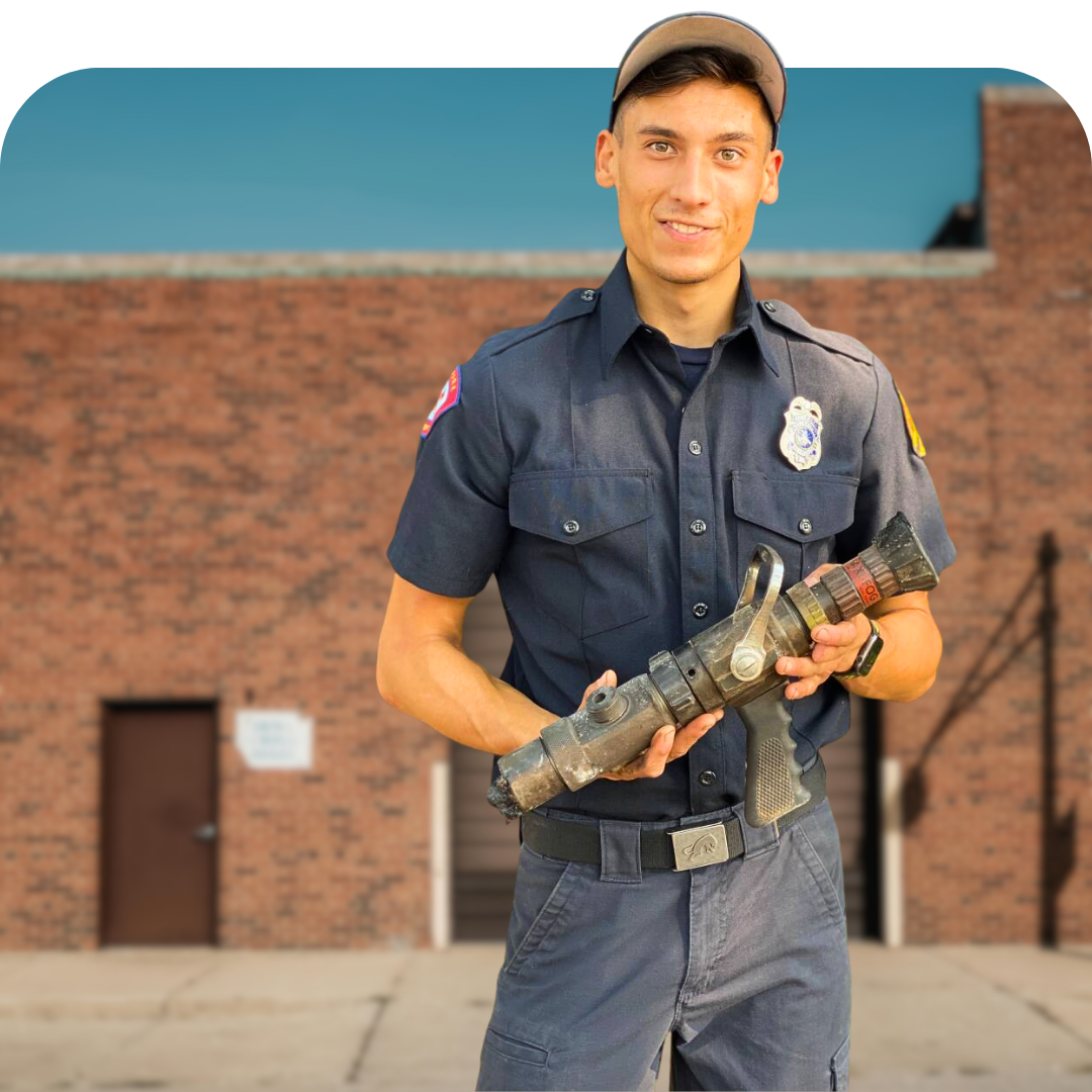 Uniformed firefighter holding a hose nozzle, standing in front of a brick building.