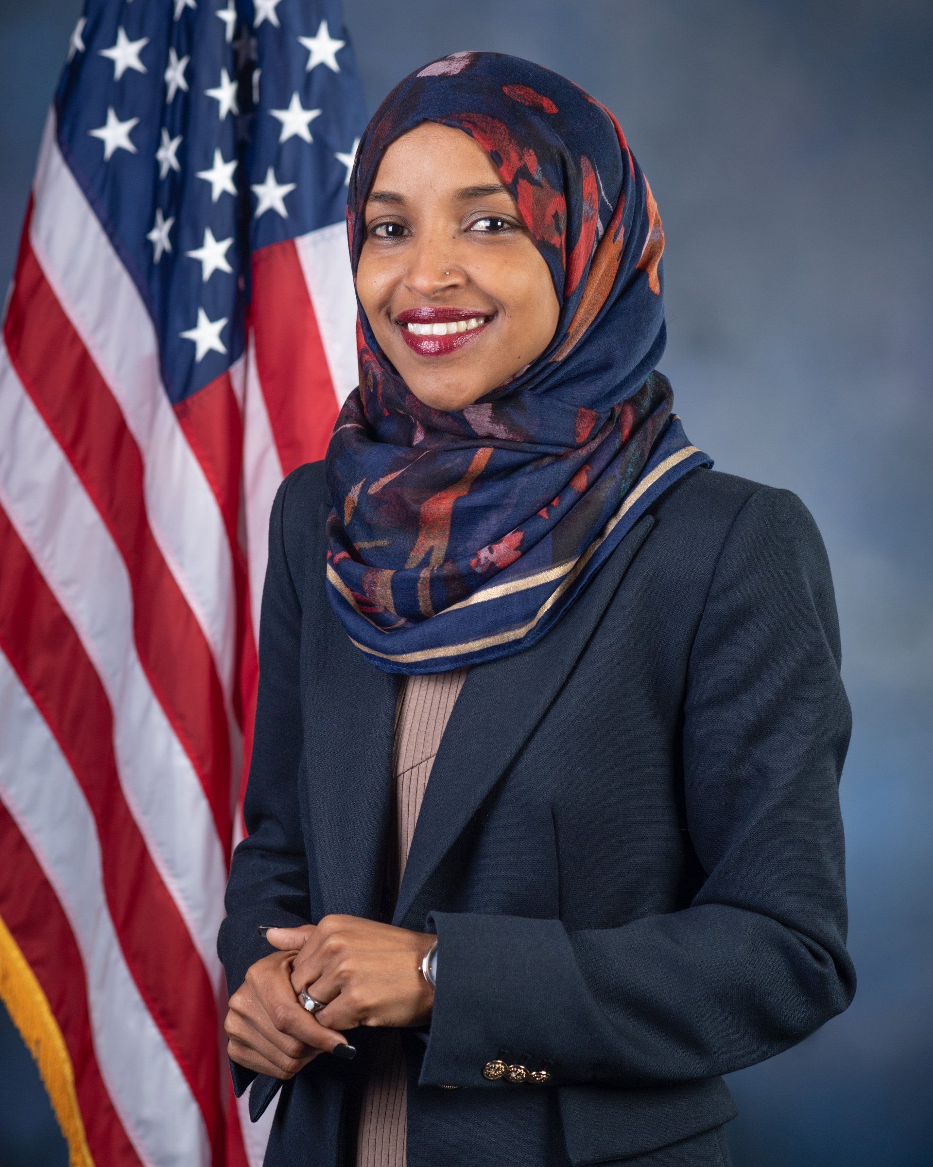 U.S. Representative Ilhan Omar in front of US flag, wearing a headscarf, and smiling.