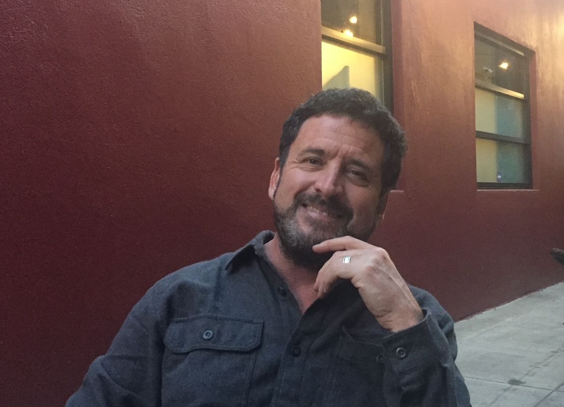 Man in a dark shirt smiles, resting a hand on his chin, sitting outside a building with a maroon wall.
