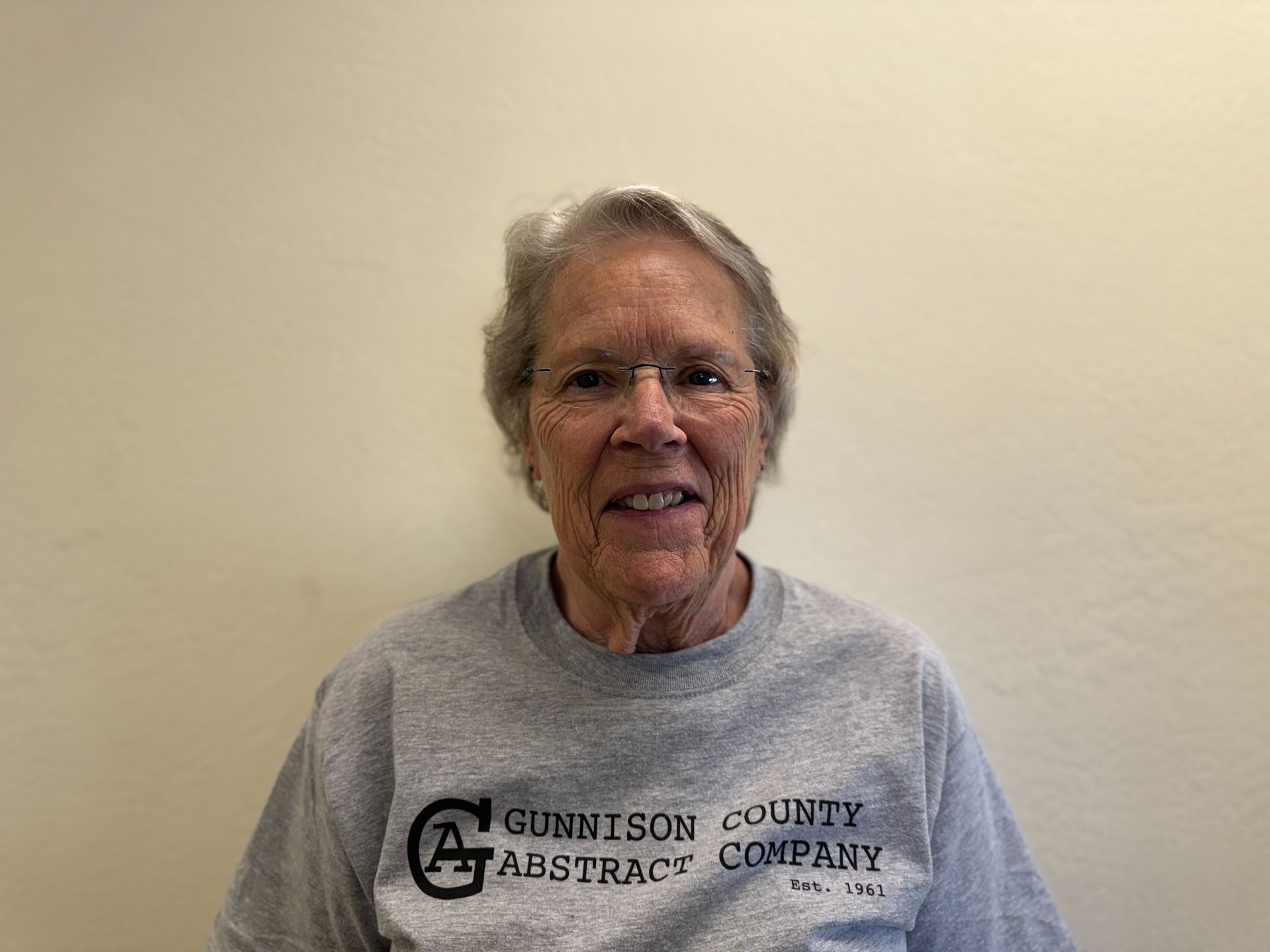 An elderly woman wearing a grey t-shirt is smiling for the camera.