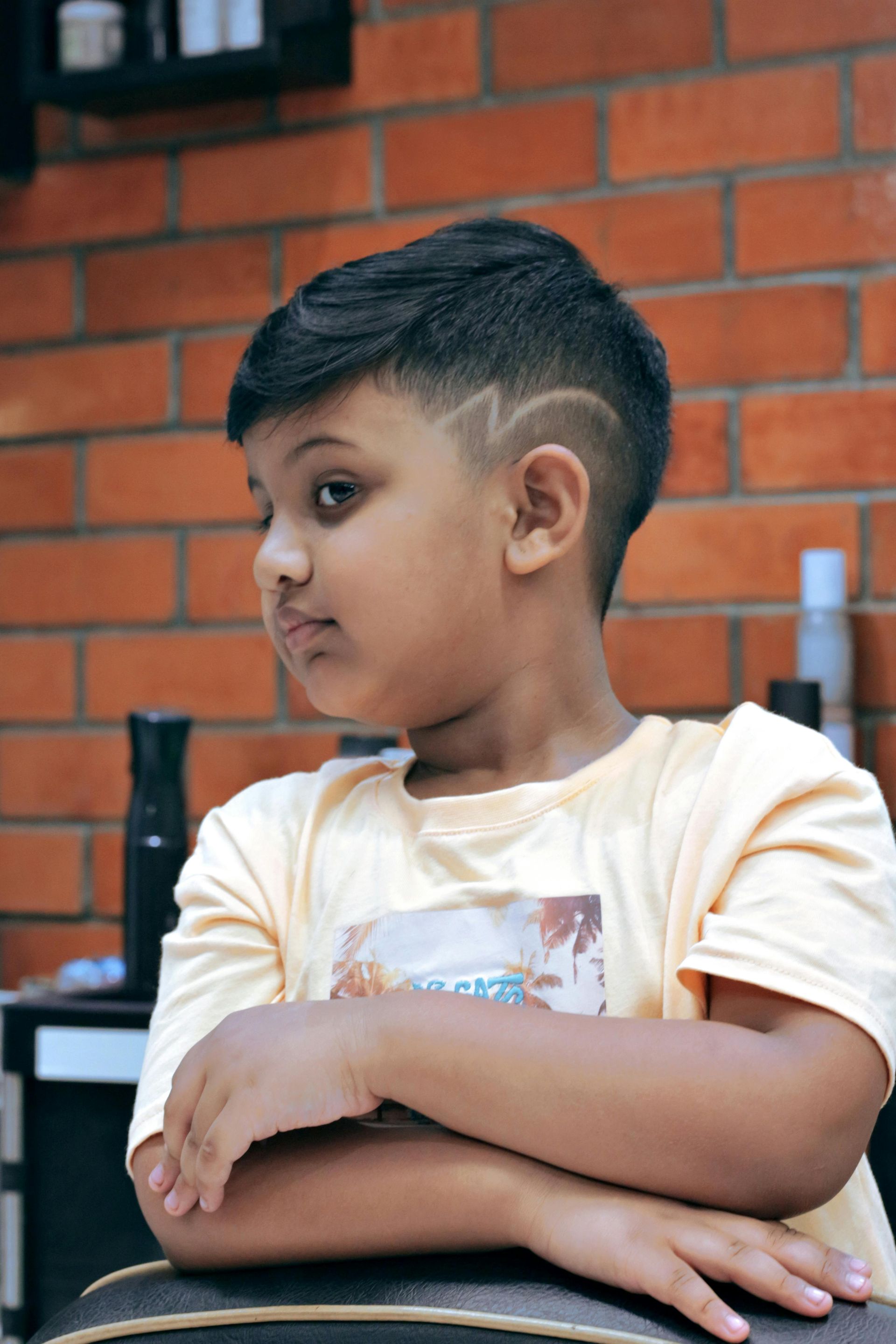 A child with a short fade haircut featuring a lightning bolt design, wearing a light yellow shirt against a brick wall.