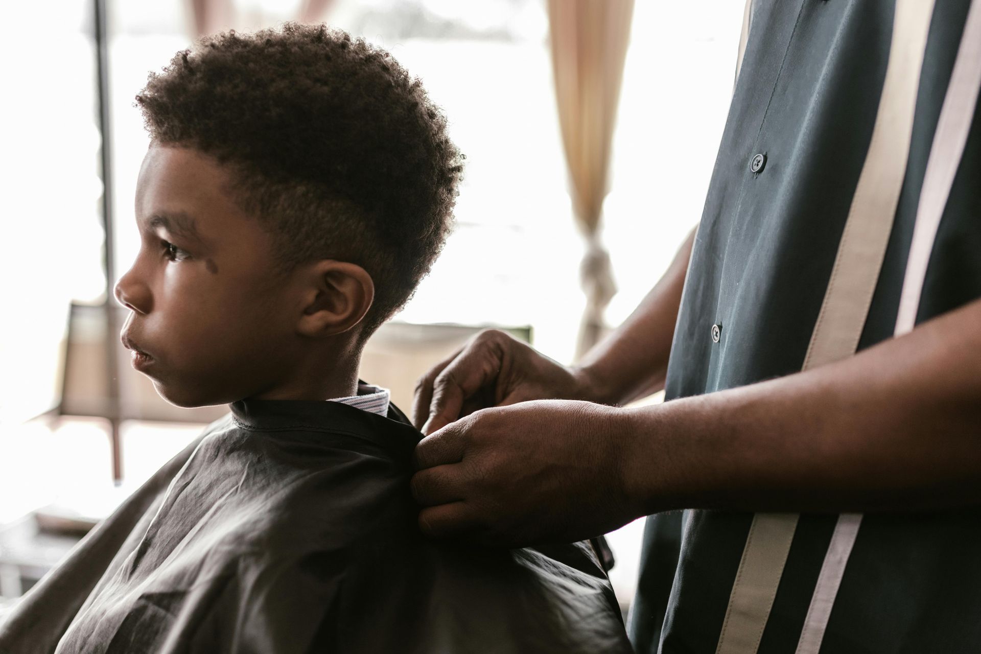 A person adjusting a barber's cape around a child's neck in a bright indoor setting.