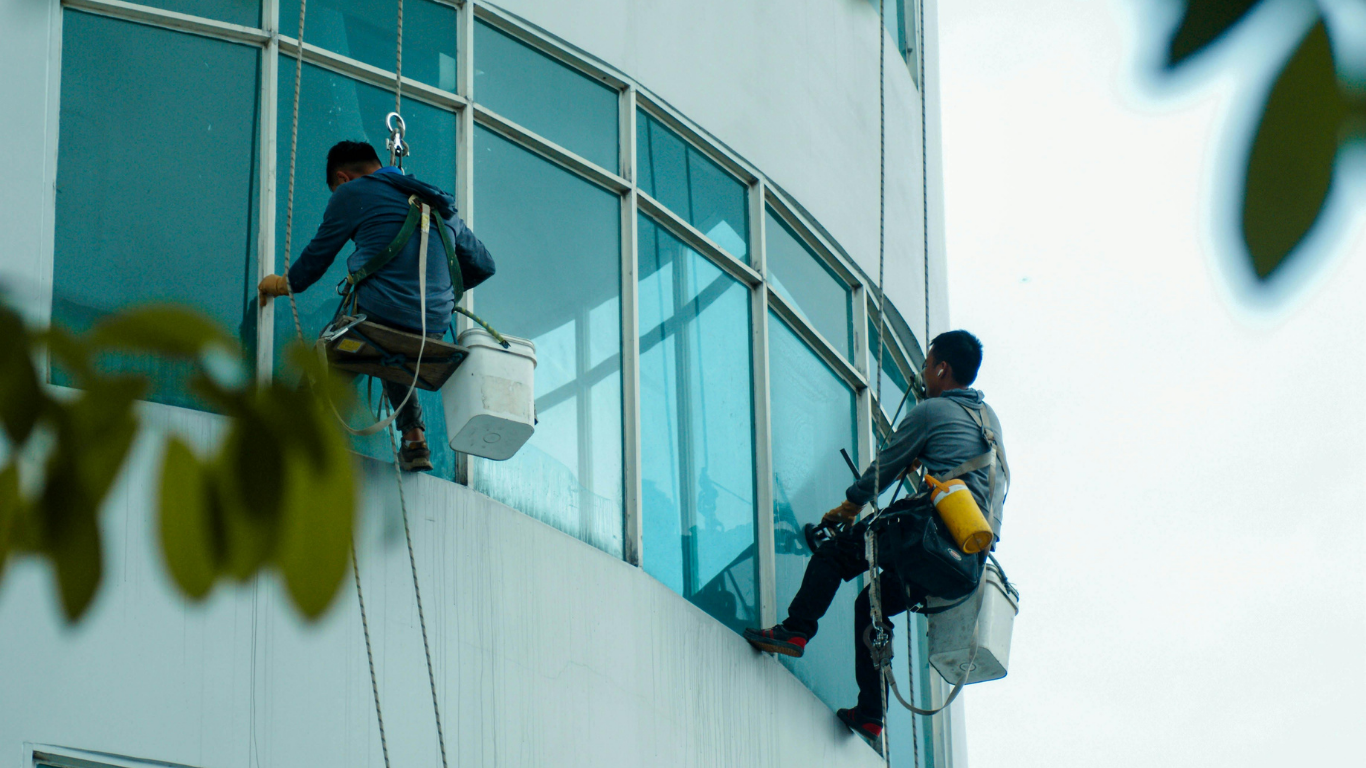 Two window cleaners suspended on ropes cleaning a curved glass building exterior.