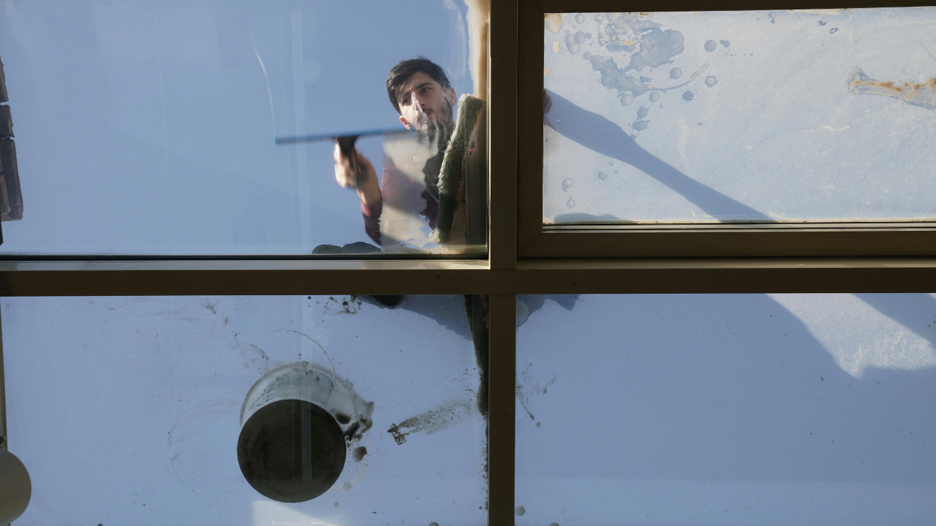 Person cleaning a window, seen through glass panes with a blue sky reflection.