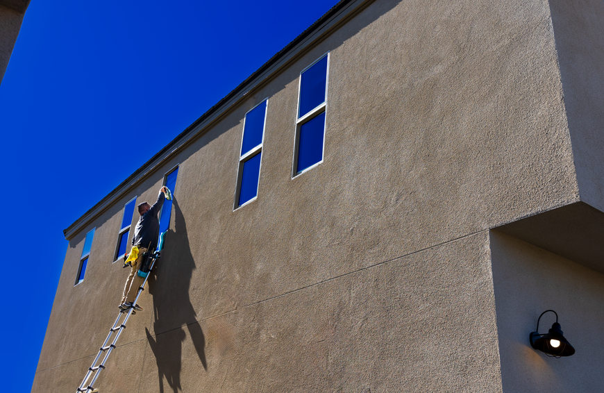 Person on a ladder painting a tan building wall near two windows under a blue sky