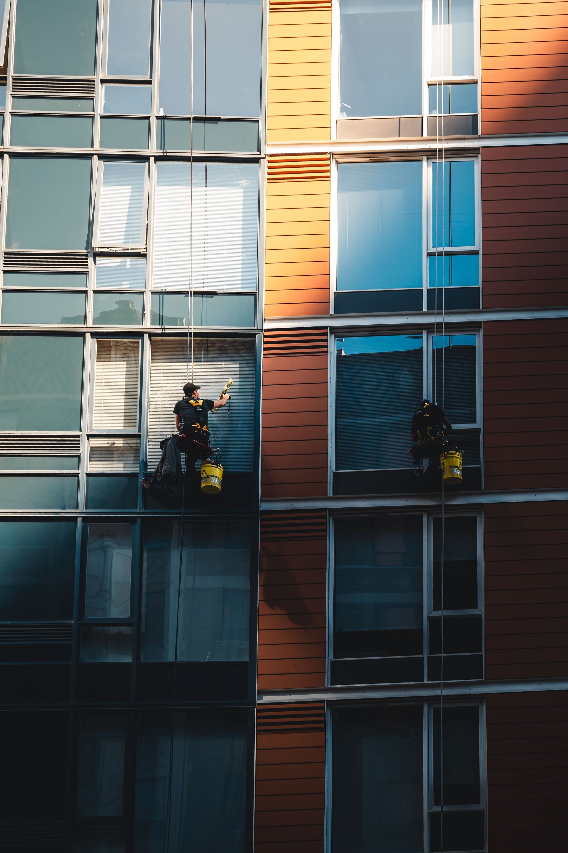 Two workers on a window cleaner’s platform beside a modern building with blue and orange panels