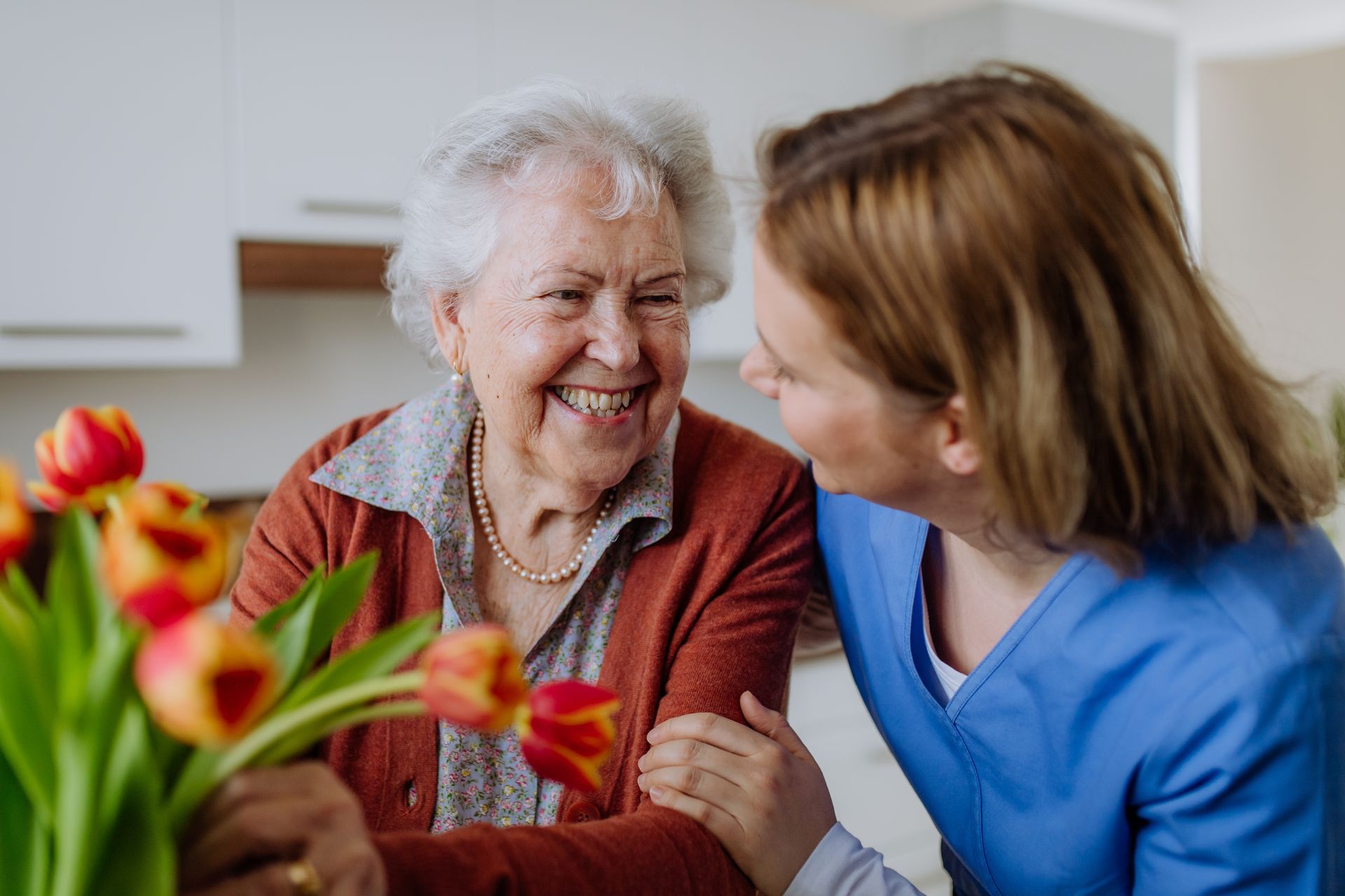 Elderly woman smiles at caregiver, holding tulips in a home setting.