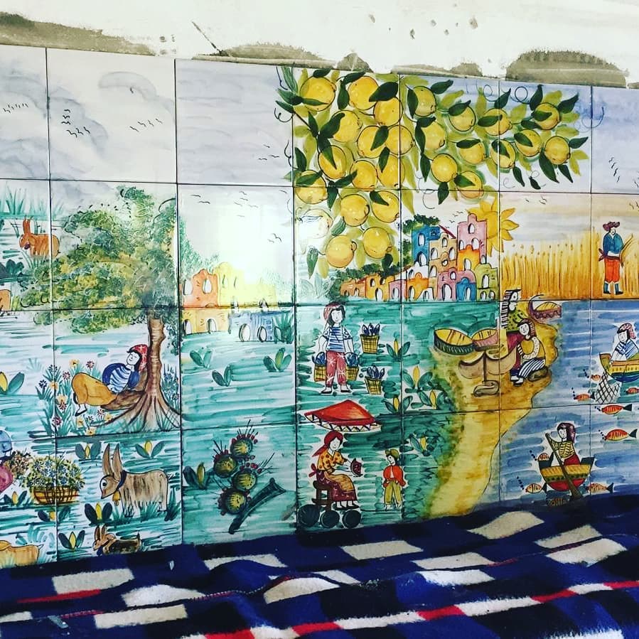 Le piastrelle dipinte a mano raffigurano scene di un giardino, una spiaggia e persone. Prevalgono i colori verde, blu e giallo.