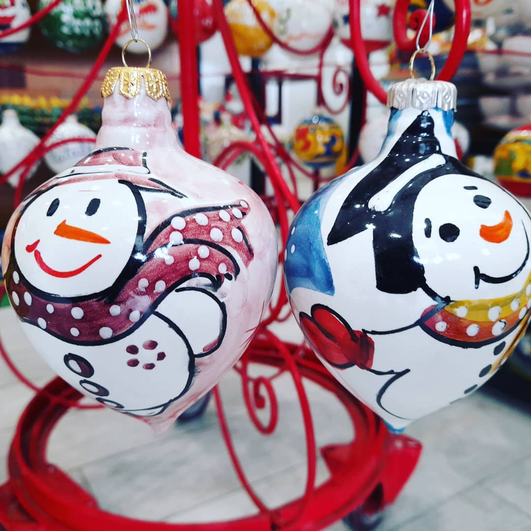 Due decorazioni a forma di pupazzo di neve, di colore rosa e blu, con volti sorridenti, appese a un supporto decorativo rosso.