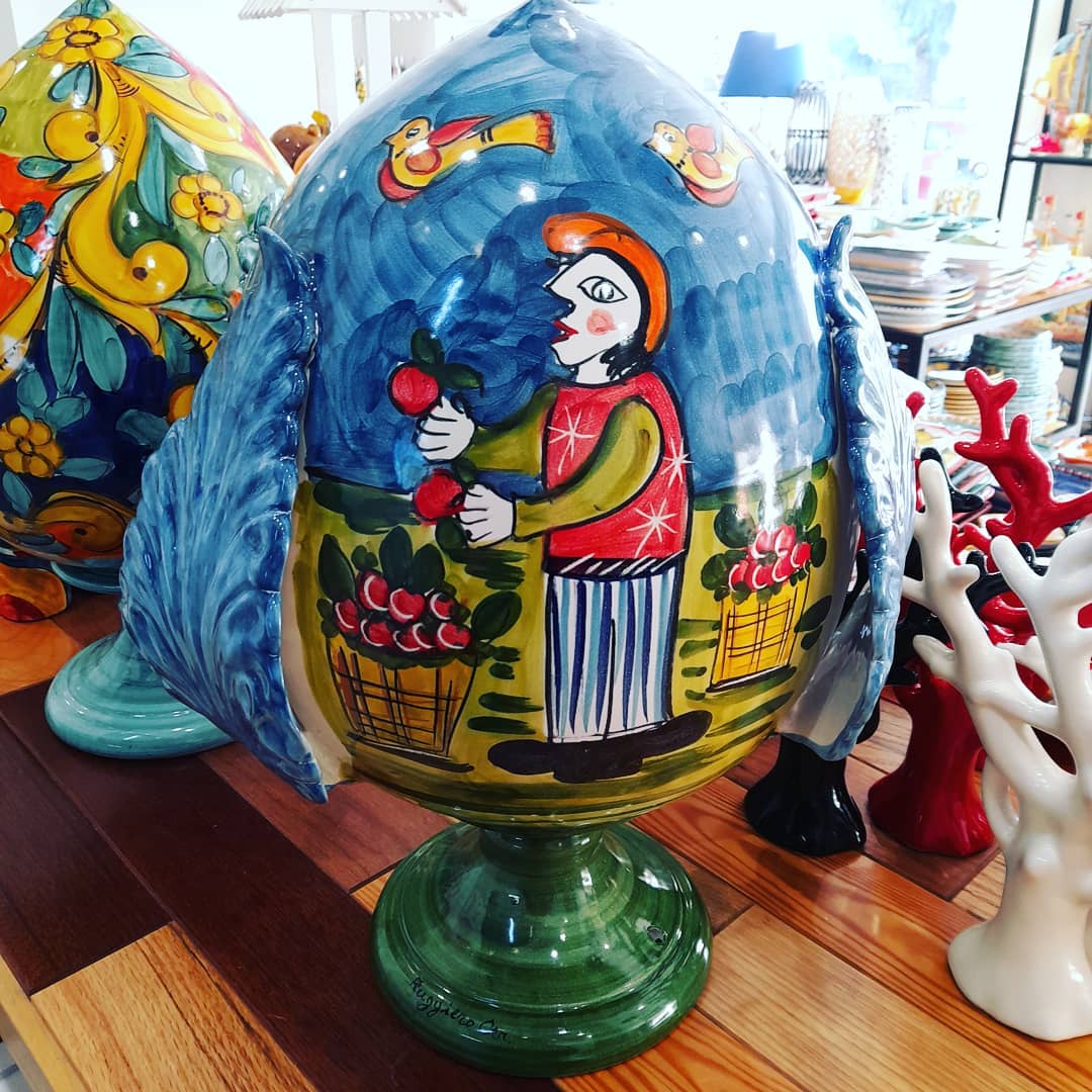 Grande scultura in ceramica colorata a forma di uovo; tonalità blu e verde, raffigurante una figura dipinta in una scena di giardino.