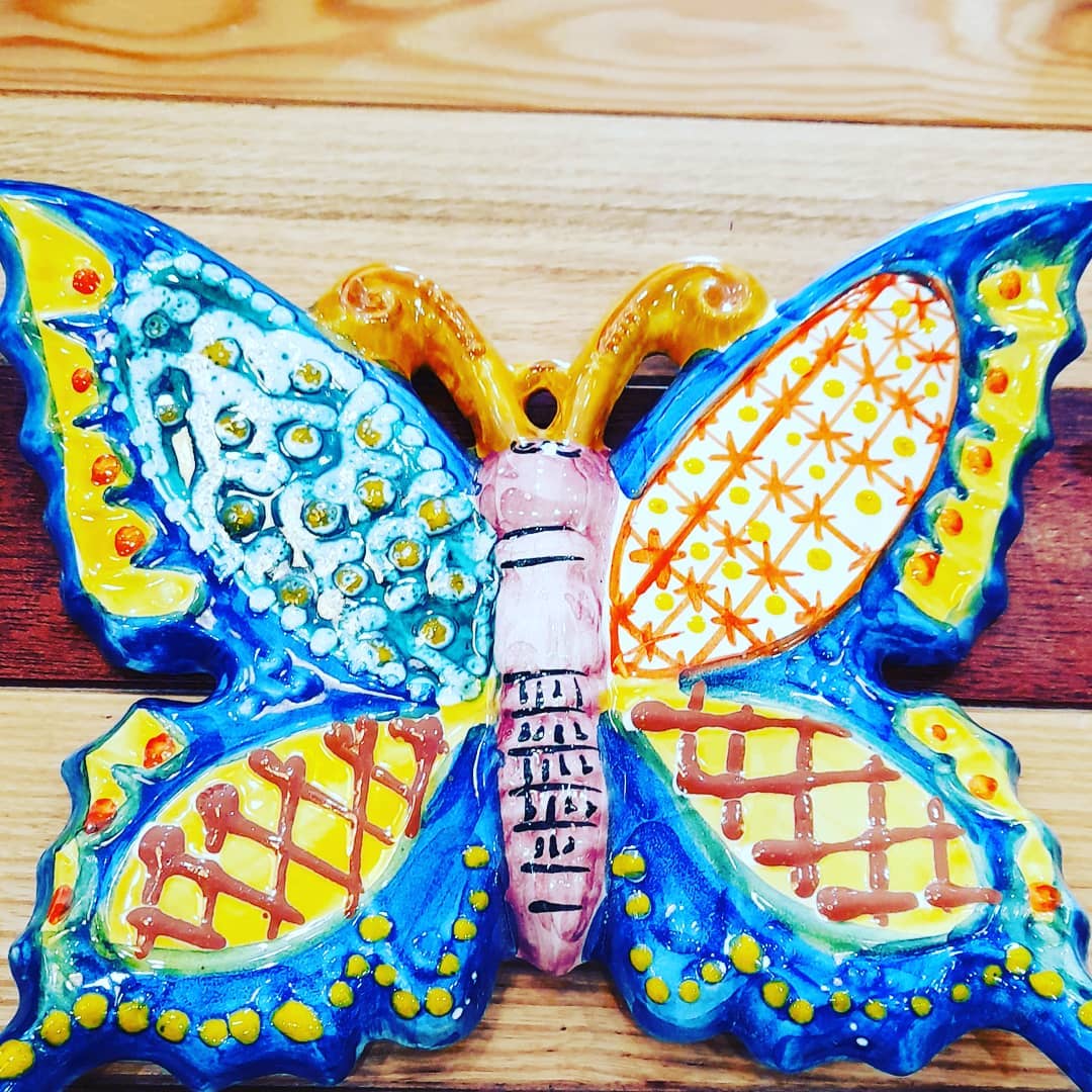 Farfalla colorata in ceramica con ali blu, gialle e arancioni, appoggiata su una superficie di legno.