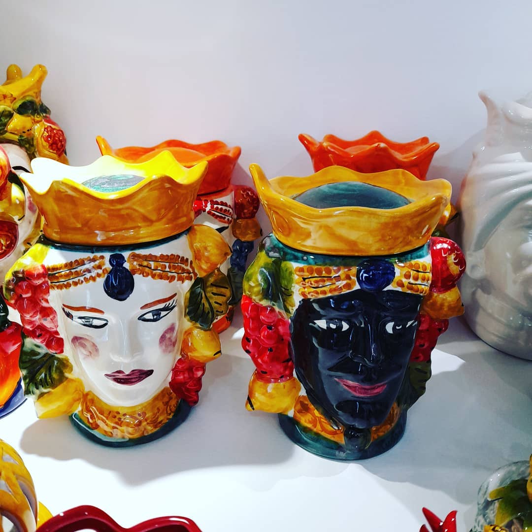 In esposizione vasi in ceramica a forma di testa, uno bianco e uno nero, con coperchi a forma di corona e dettagli colorati.