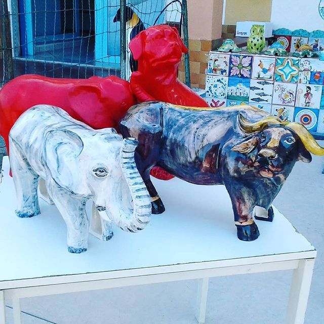 Sculture di animali colorati su un tavolo bianco: cane rosso, mucca rossa, elefante grigio e toro nero.