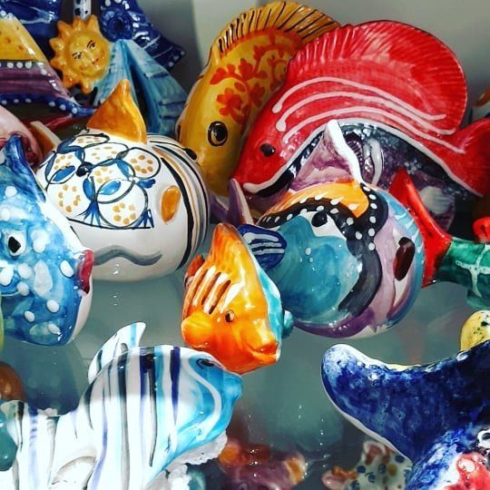 Sculture di pesci in ceramica colorata, primo piano. Varie dimensioni e motivi nei colori blu, rosso, arancione, giallo.