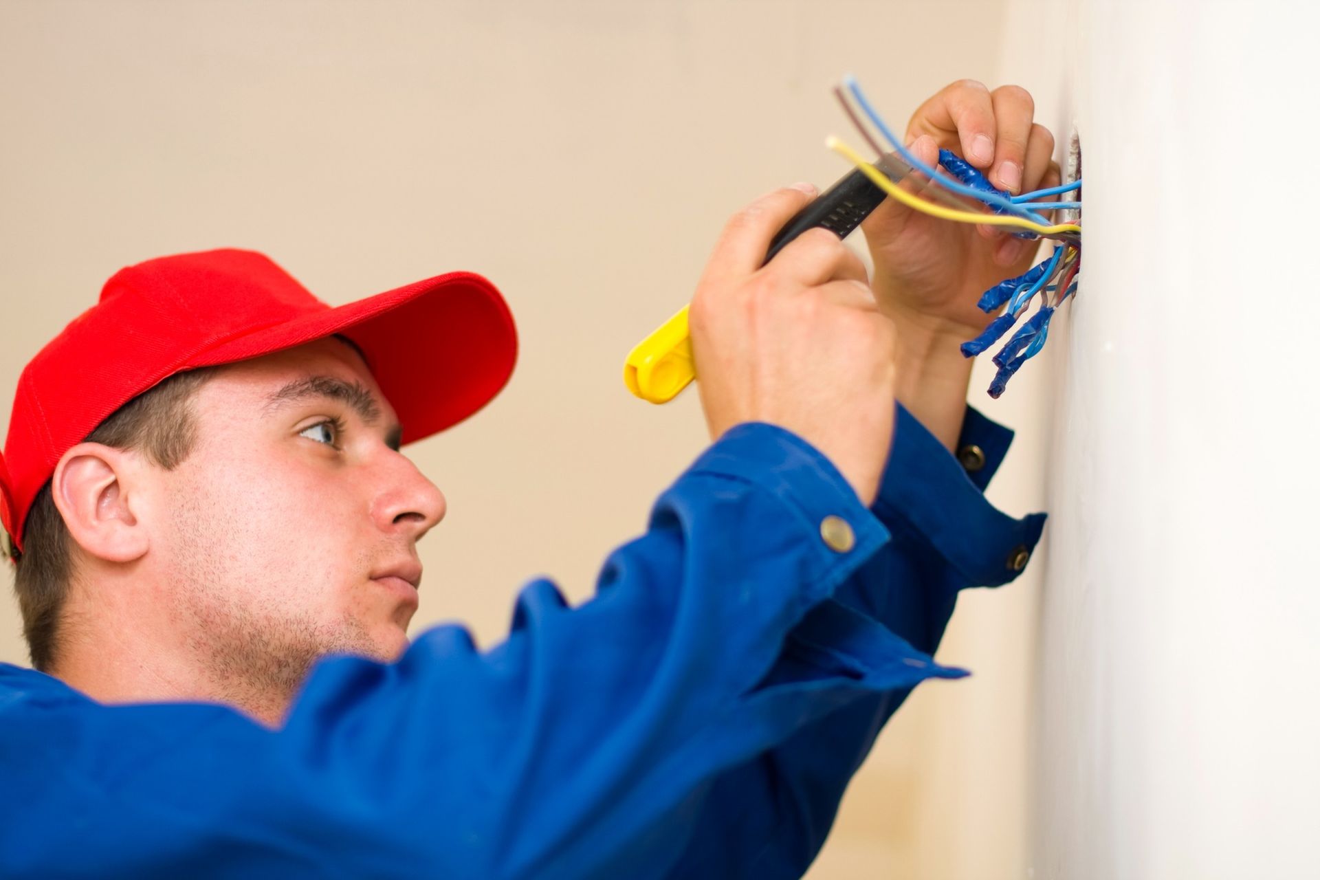 Electrician in blue coveralls and red cap working on wires in a wall outlet.