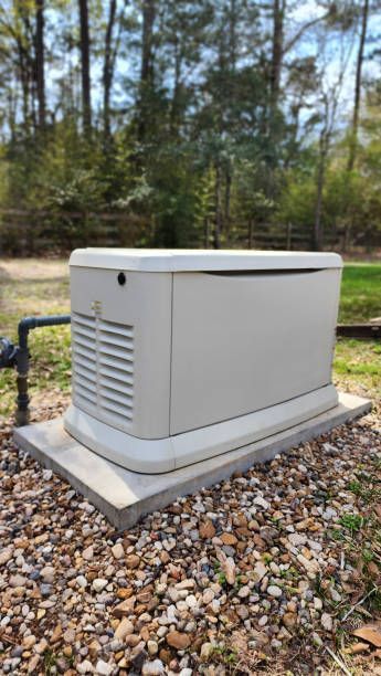 A light-colored standby generator on a concrete pad, gravel ground, wooded background.