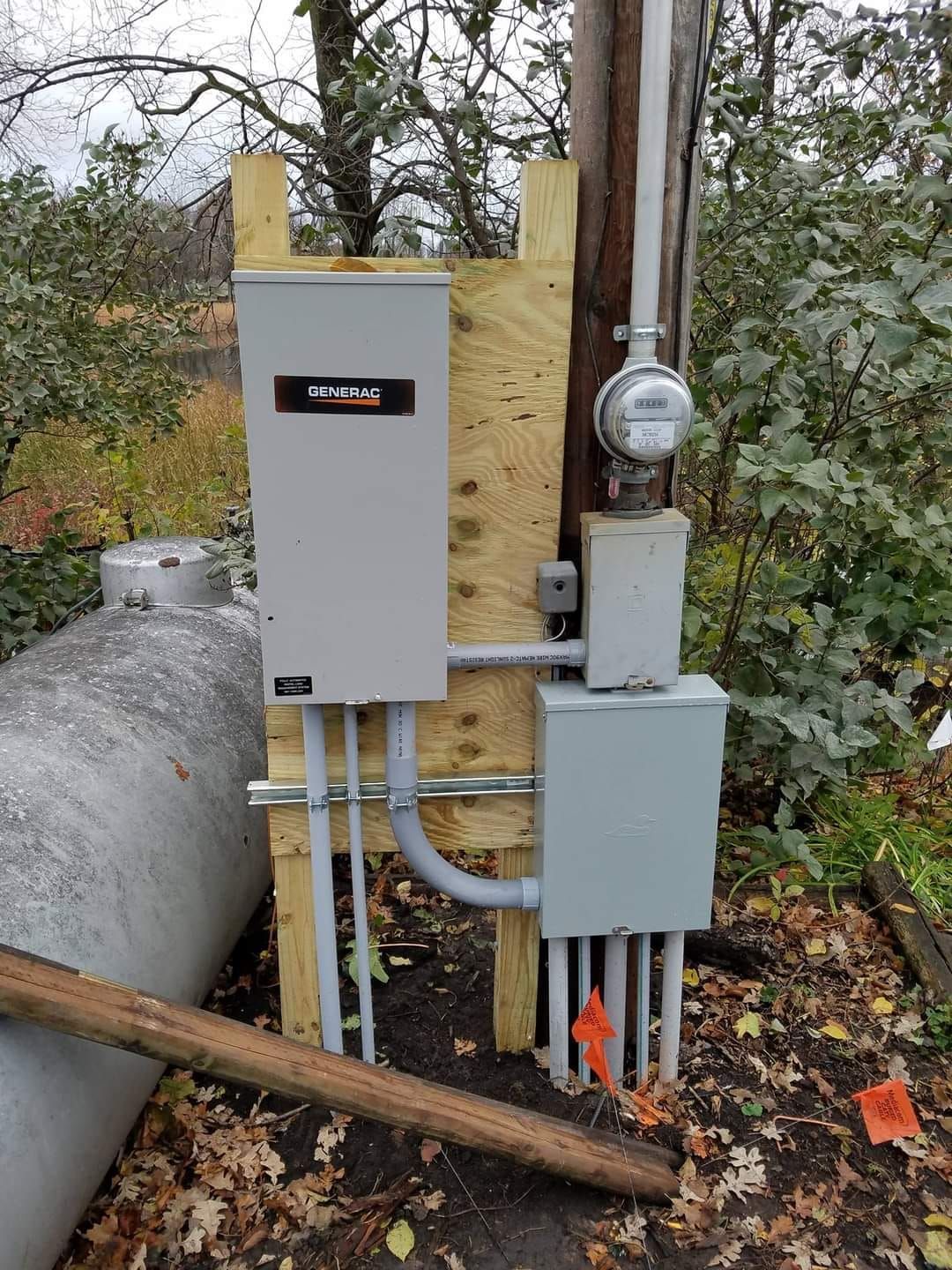 Electrical panel and meter mounted on wood, near a propane tank and utility pole outdoors.