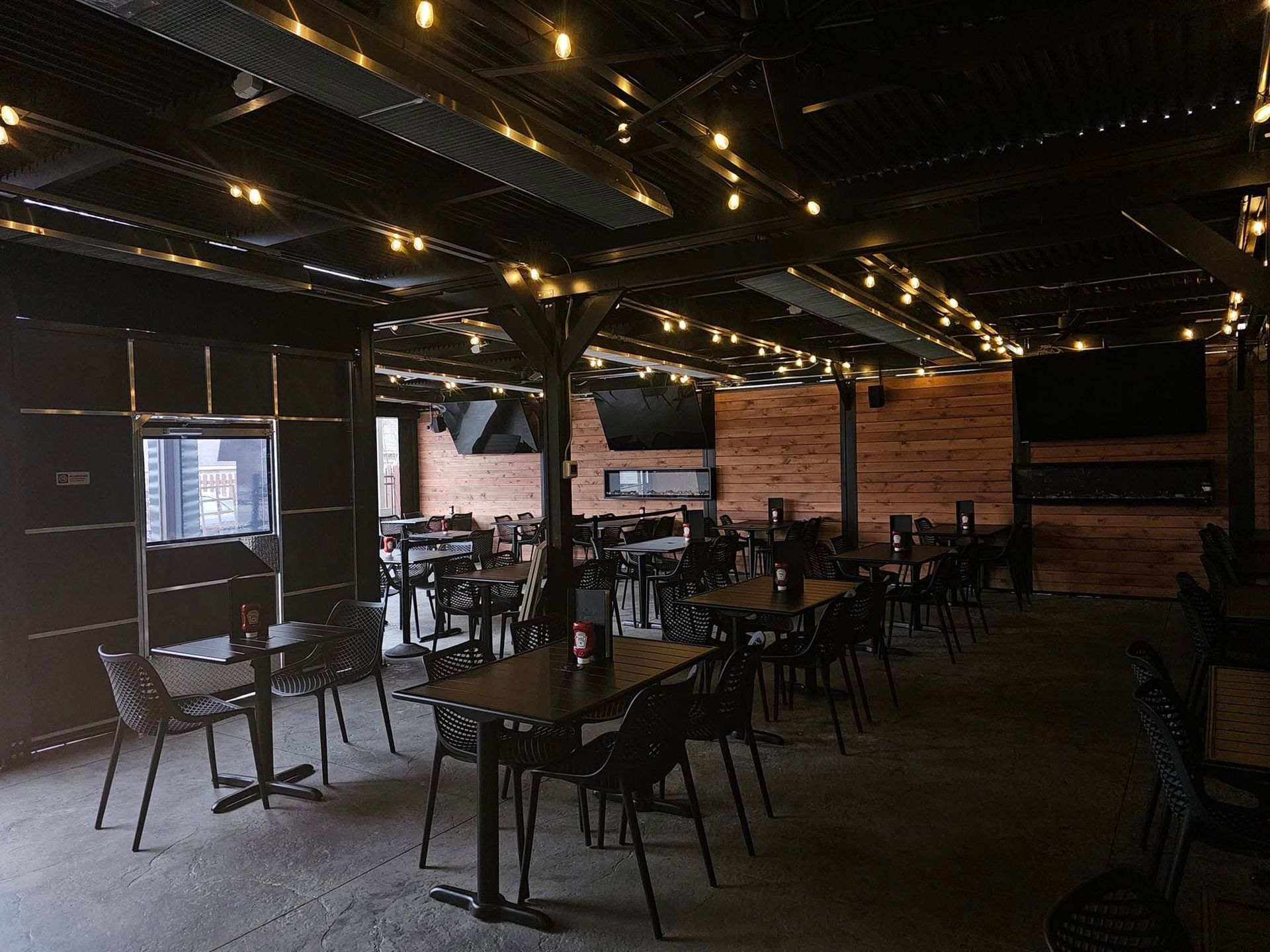 Outdoor restaurant patio with tables, chairs, string lights, and a wooden wall.