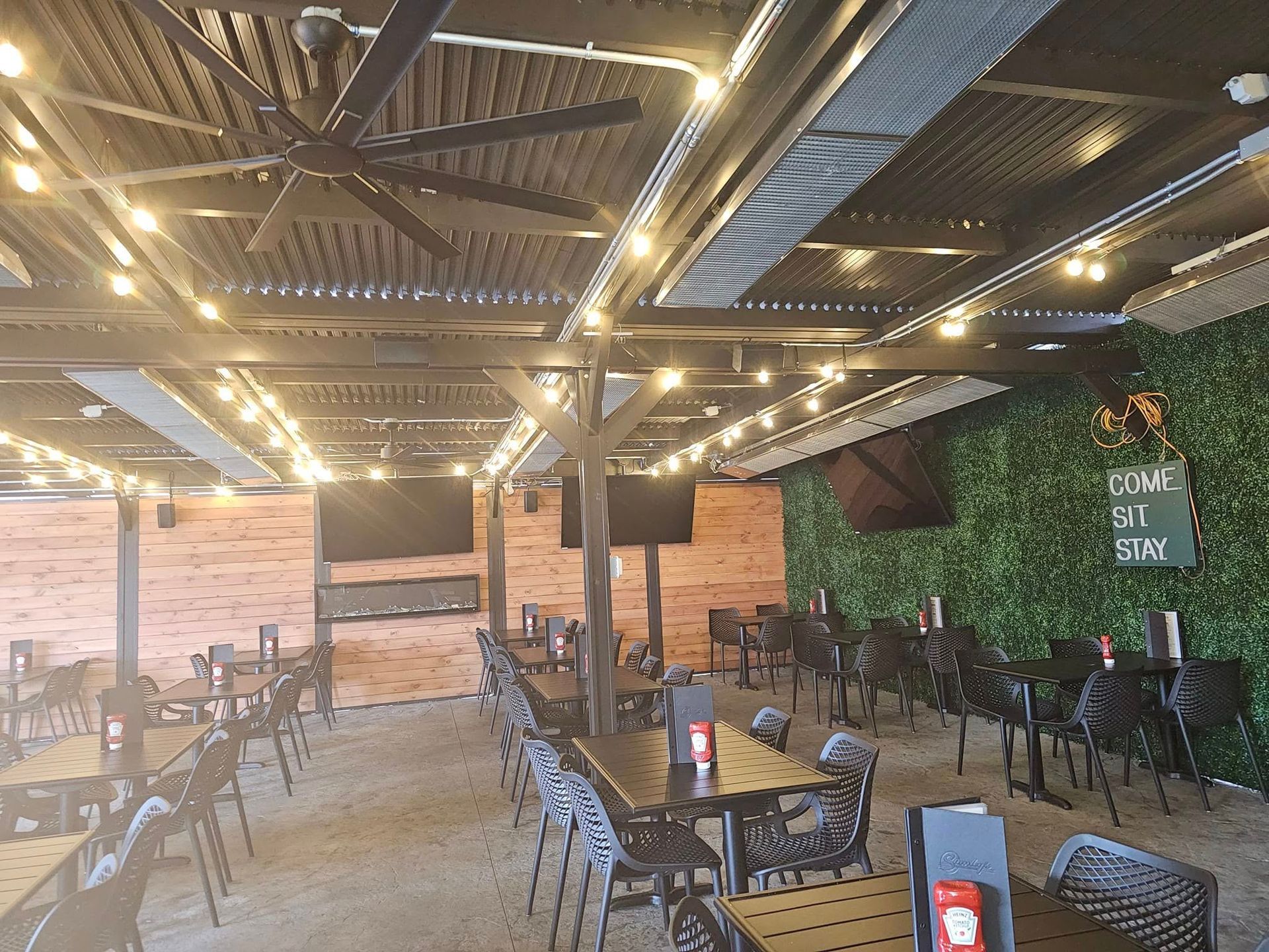 Outdoor patio with tables, chairs, TVs, string lights, and a faux-greenery wall.