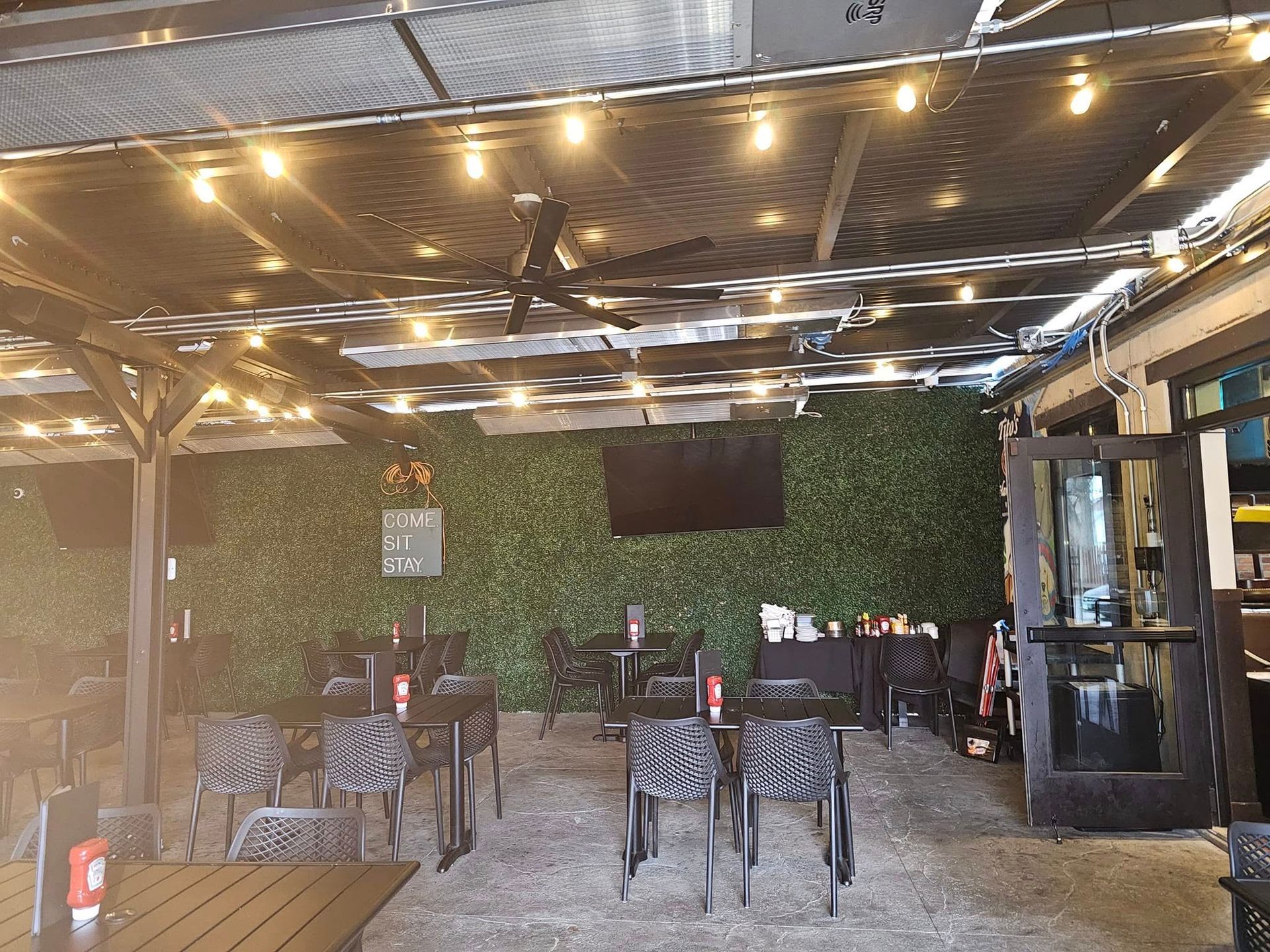 Outdoor patio with string lights, black tables and chairs, a green hedge wall, and a TV screen.
