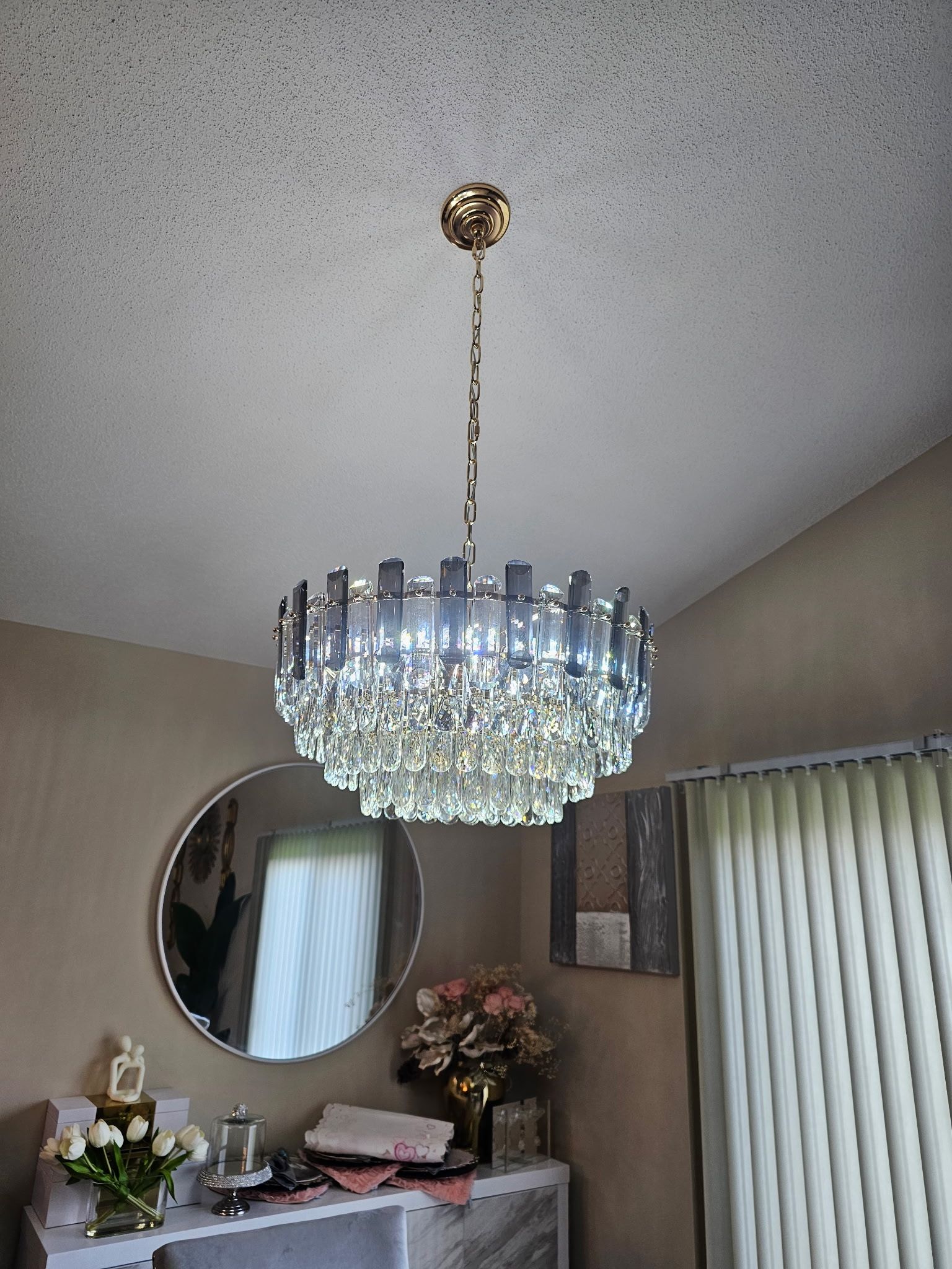 Crystal chandelier hanging from a gold ceiling mount. Beige walls and a round mirror are in view.