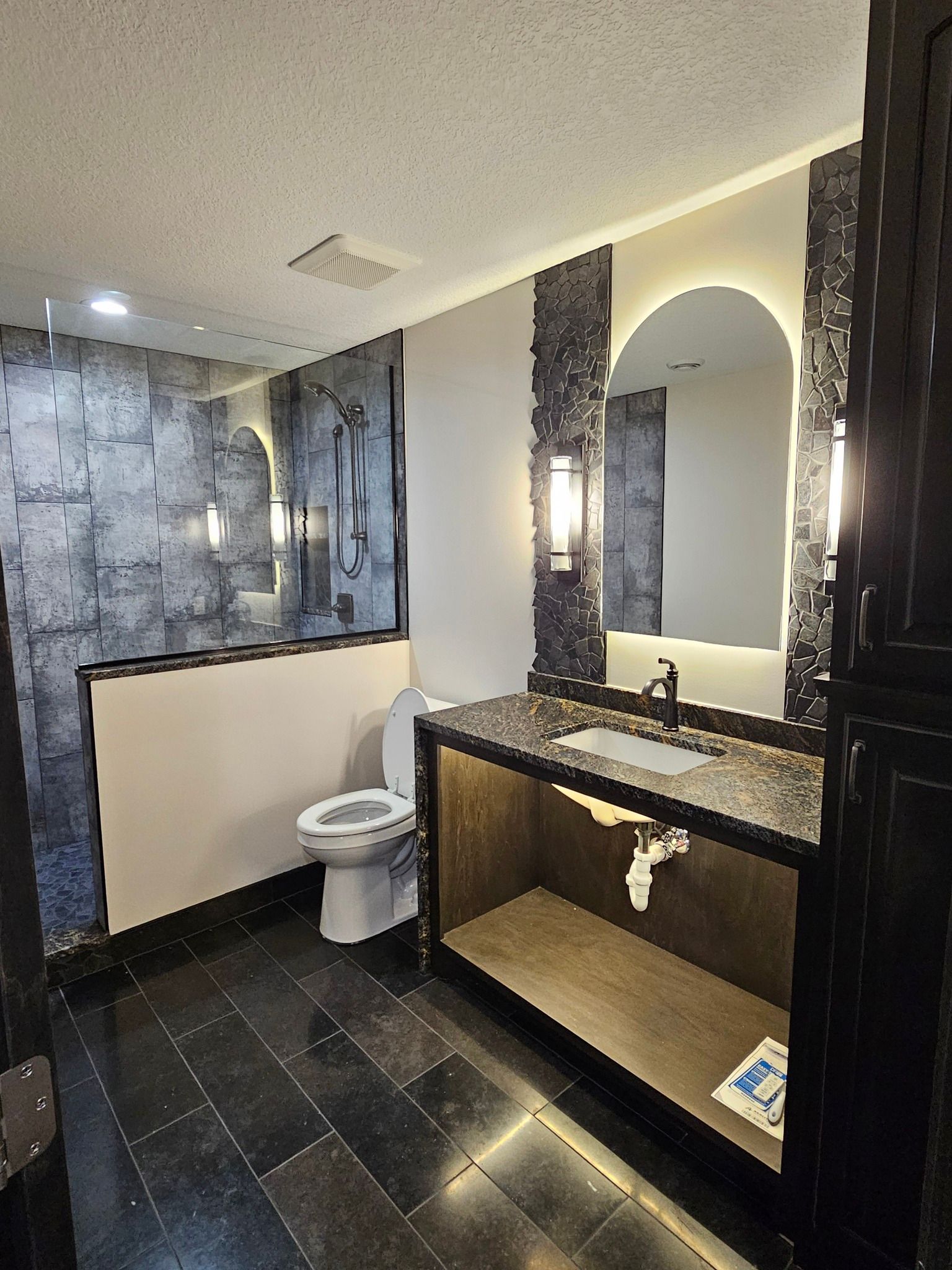 Modern bathroom with dark floor, shower, toilet, vanity, and large arched mirror.