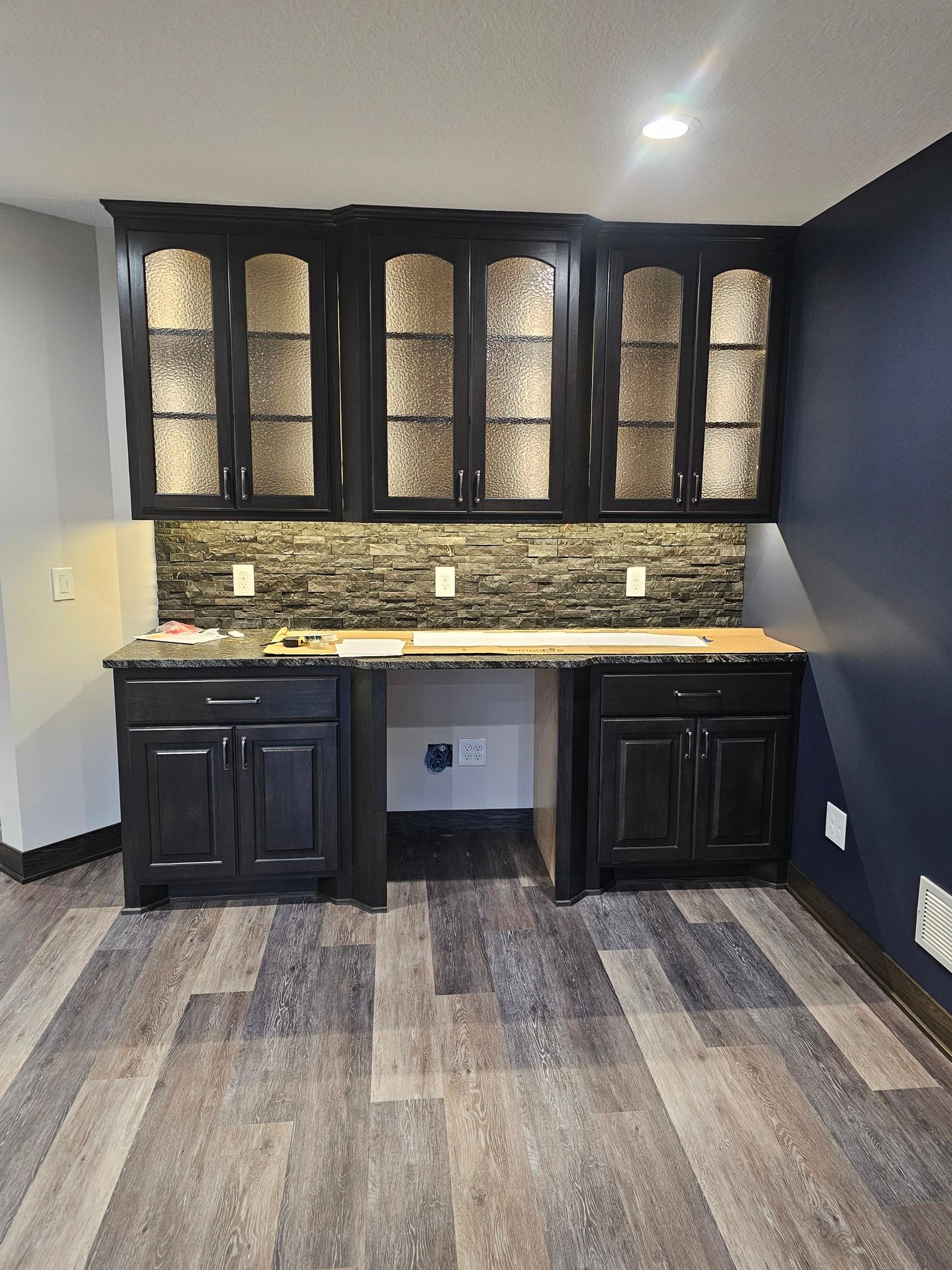 Black built-in desk with glass-door cabinets, tiled backsplash, and wooden countertop, against blue and gray walls.