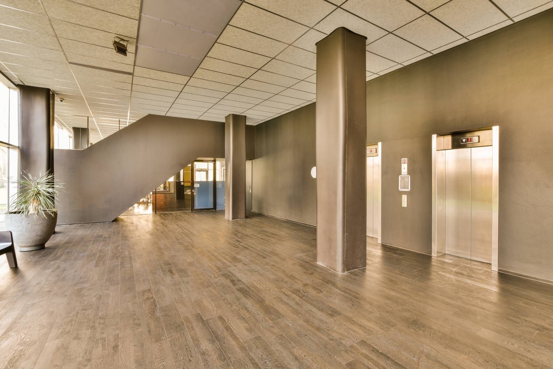 Empty modern lobby with wood floors, stairs, elevator, and tall windows