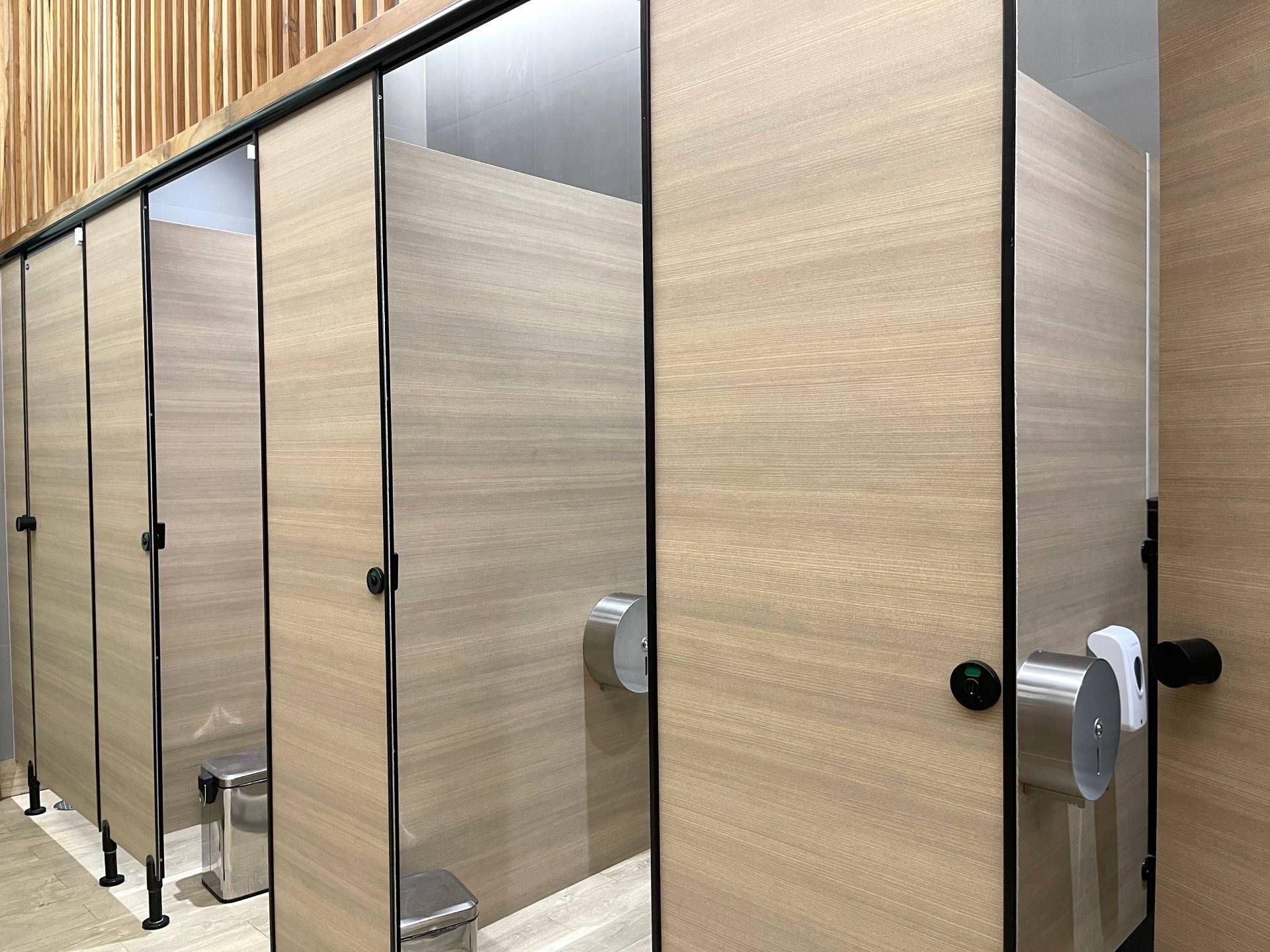 Row of wood-finish restroom stall doors in a public bathroom with tiled floor and grab bars