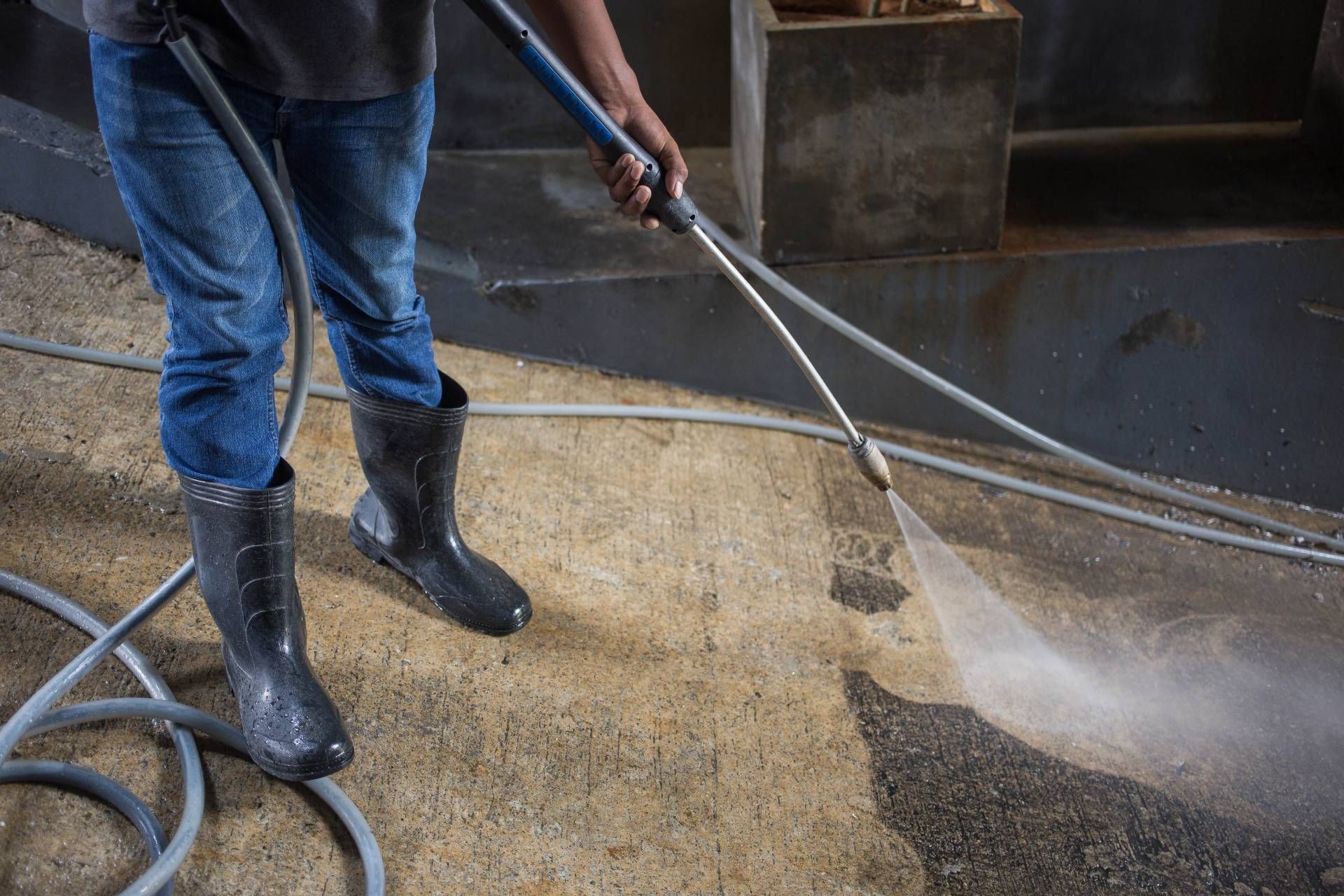 Person pressure-washes a wet concrete floor with a hose and wand in black boots.