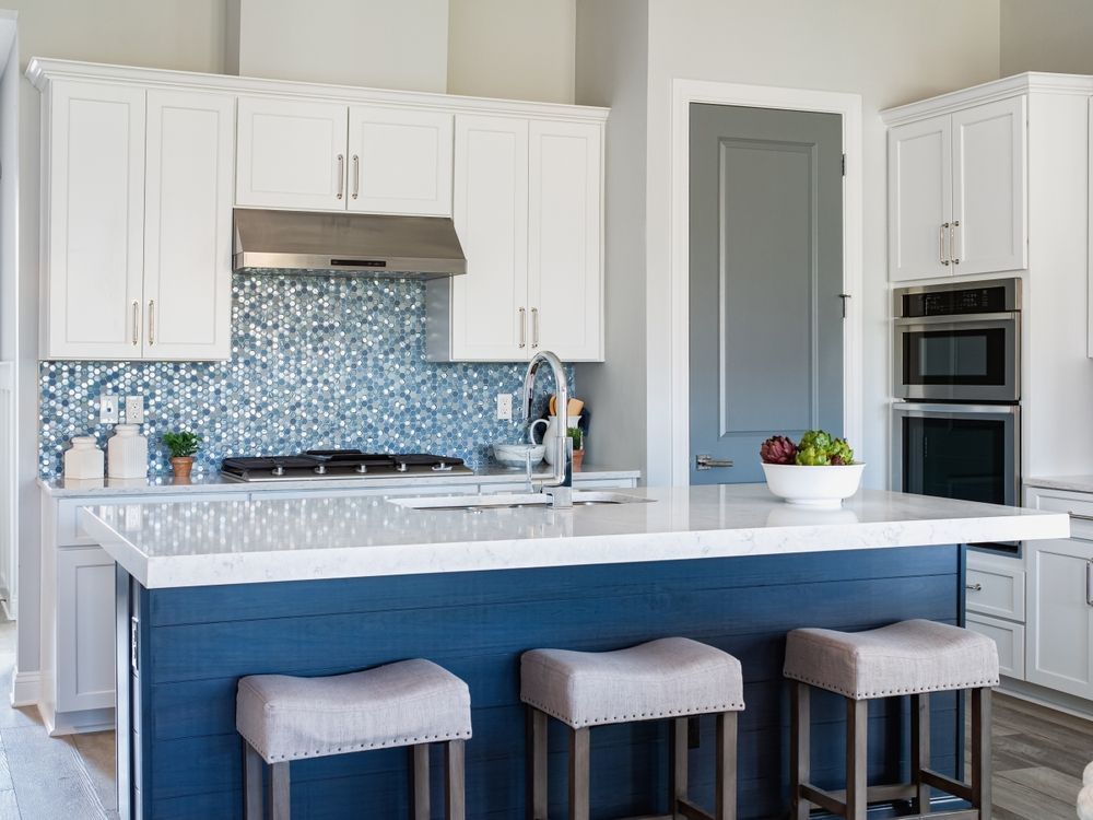 Modern white kitchen with blue island, three stools, and built-in oven