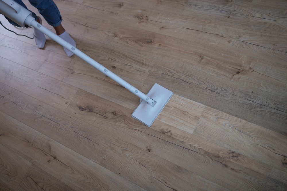 Person vacuuming a light wood floor with a white stick vacuum