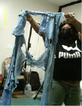 Person holding up heavily ripped denim pants. Black Puma shirt, mask, room setting.