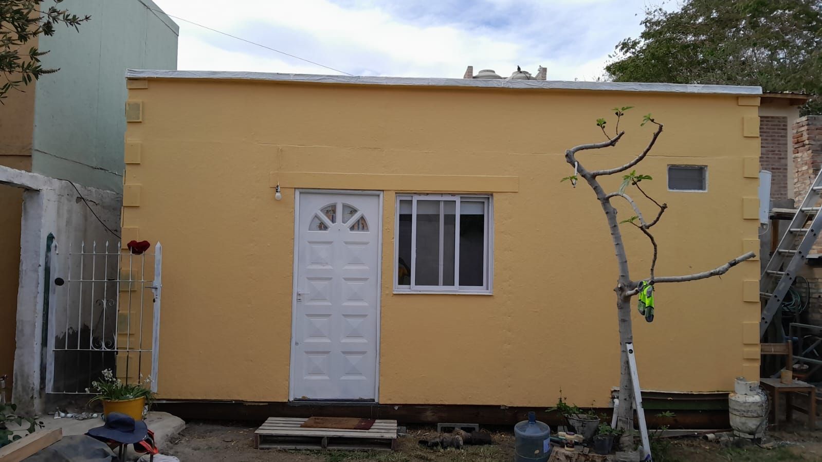 Edificio rectangular amarillo con puerta y ventana blancas; un árbol desnudo a la derecha.