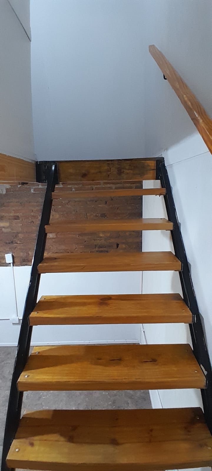 Escalera de madera con estructura de metal negro que sube hasta una pared de ladrillo y una pared pintada de blanco.