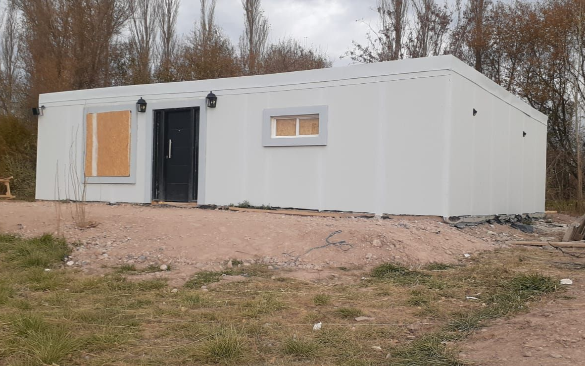 Edificio rectangular blanco con puerta negra y dos ventanas, situado en una zona verde con árboles al fondo.